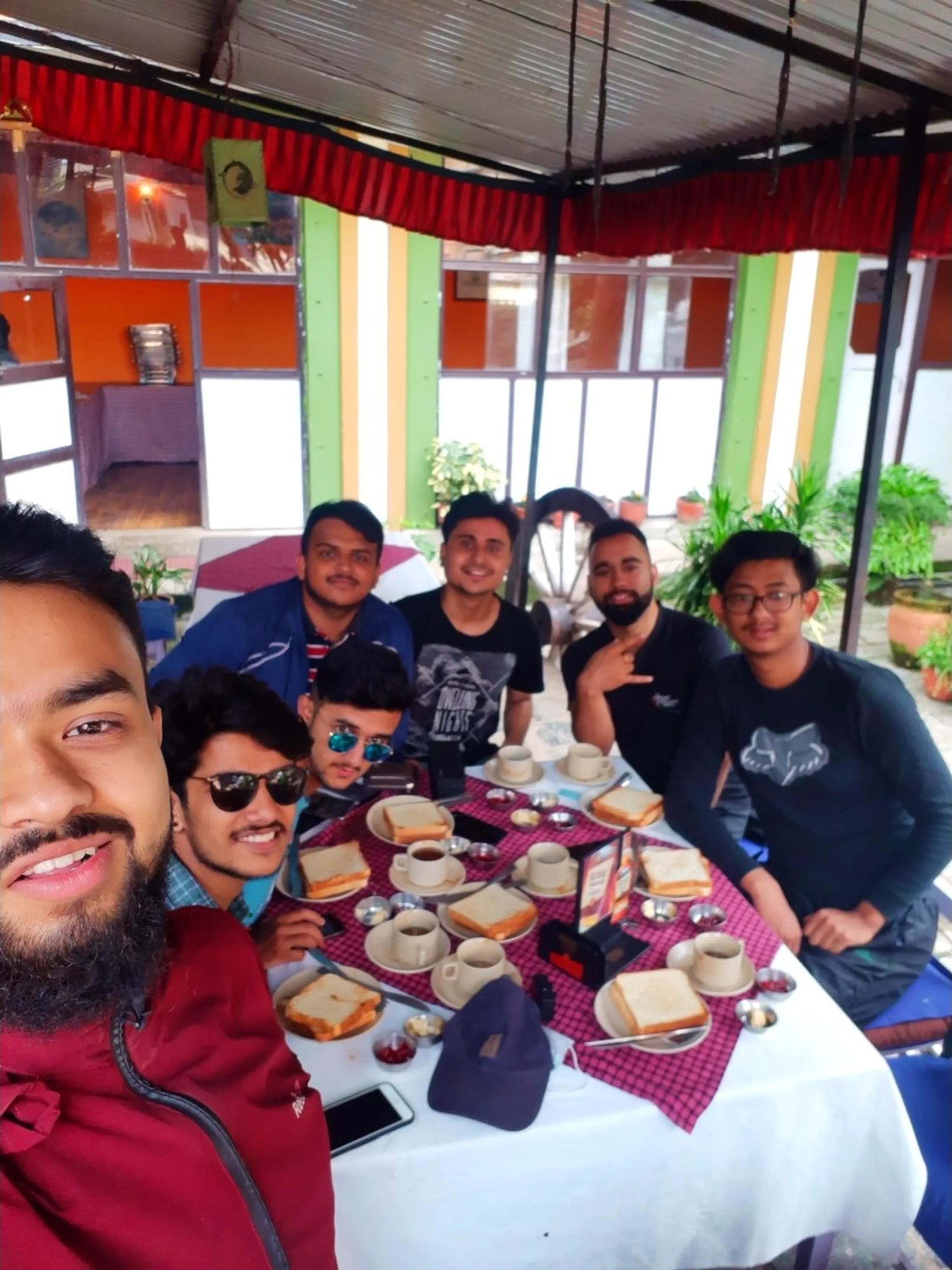 Group of seven young Nepali male friends posing together for a wide-angle selfie at an outdoor restaurant terrace before starting their Bethanchowk hiking adventure near Kathmandu, Nepal. The photo is taken from below by a bearded man in a maroon hoodie holding the camera close, with six other friends crouched and standing behind a table loaded with breakfast spread including bread toast slices, boiled eggs in white bowls, tea cups and saucers, small steel bowls of pickles and condiments, smartphones, and a navy blue cap. The setting has a red-curtained tin roof shed, green and orange painted pillars, potted plants, and a building with orange and white walls in the background. Two friends in front wear sunglasses, others wear casual t-shirts and jackets in dark blue, grey, and black. This breakfast stop captures the excitement before a Nepal motorbike trekking trip to Bethanchowk, Kathmandu Valley adventure, group hiking trip Nepal, friends road trip Nepal travel blog.