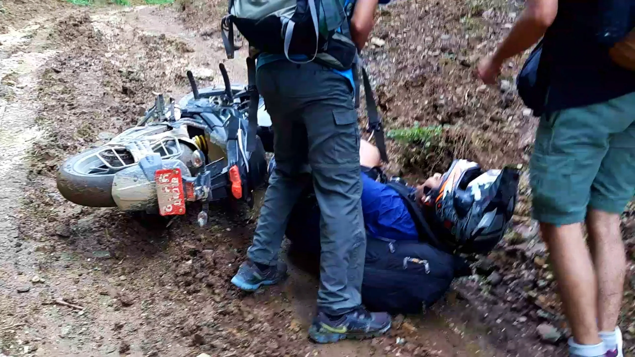 Aftermath photo of a motorbike accident on a muddy forest trail in Nepal, showing a black motorcycle tipped over on its side in thick mud with a red Nepali license plate visible on the rear, wheels and undercarriage caked in brown mud. A person lies on the ground wearing a blue jacket with a black backpack and motorcycle helmet, being checked on by another hiker standing over them wearing grey cargo pants, hiking boots, and a backpack with sleeping mat. A third person in green shorts and a dark top stands nearby on the right side of the frame. The muddy dirt trail is flanked by eroded reddish soil banks and green vegetation. This documents a real motorbike trekking accident on Bethanchowk trail, off-road riding hazards Nepal, adventure trekking risks Himalayas, group hiking safety Kathmandu hills.