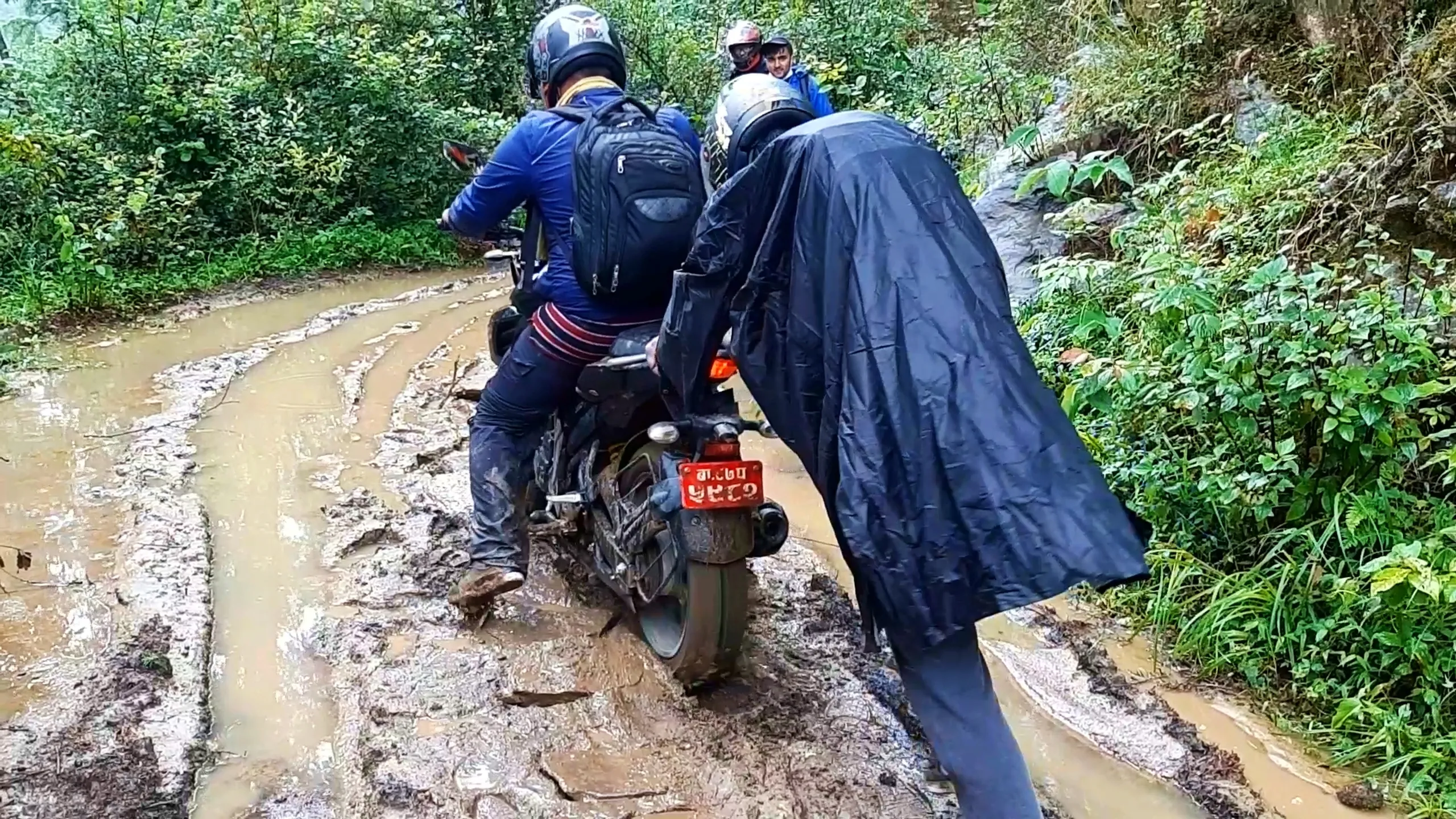 Action photo of two motorcyclists wearing helmets and rain gear riding through a deep muddy water-filled trail in a dense forest, with the rider in front wearing a blue jacket and grey backpack on a black motorbike with red Nepali number plate, while another hiker wearing a black rain poncho leans over the bike's rear from behind. In the background, two more figures wearing helmets watch from the muddy path, surrounded by lush green forest vegetation on both sides. This captures the challenging muddy stream crossing during Bethanchowk motorbike trekking, Nepal monsoon off-road riding, Himalayan flooded trail adventure, group motorbike trekking Kathmandu hills.