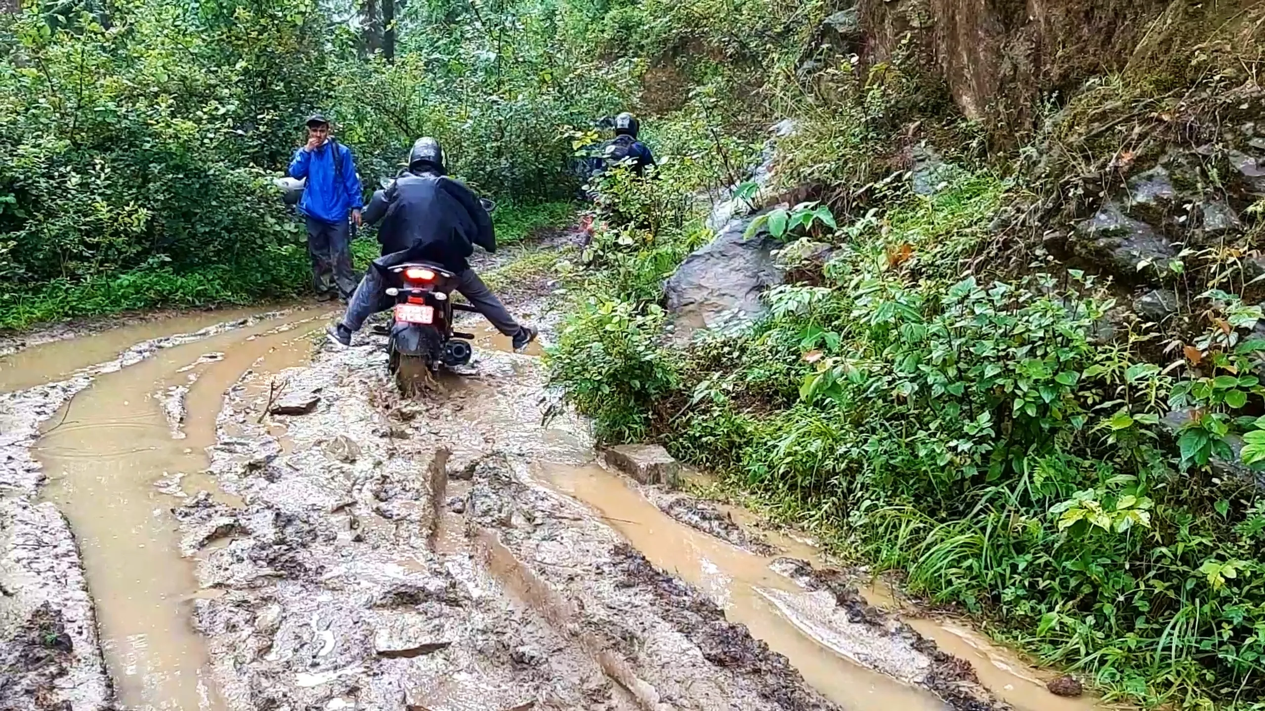 A muddy forest trail in Bethanchowk, Kathmandu Valley, Nepal, churned into thick brown mud by rain during a monsoon motorbike trek. A rider in a black raincoat and helmet straddles a motorcycle stuck in a deep muddy puddle, both feet planted on the ground for balance, rear wheel splattered with mud, red taillight glowing. Ahead on the trail, another rider in a black helmet and backpack walks alongside his bike. To the left, a man in a blue rain jacket and cap holds a helmet, watching. Dense green forest, ferns, and a rocky moss-covered hillside line the narrow dirt track, with muddy water channels running downhill. Ideal for Nepal trekking blog, Himalayan adventure travel, Bethanchowk hiking trail, off-road motorbike ride Nepal, monsoon trekking Kathmandu Valley.