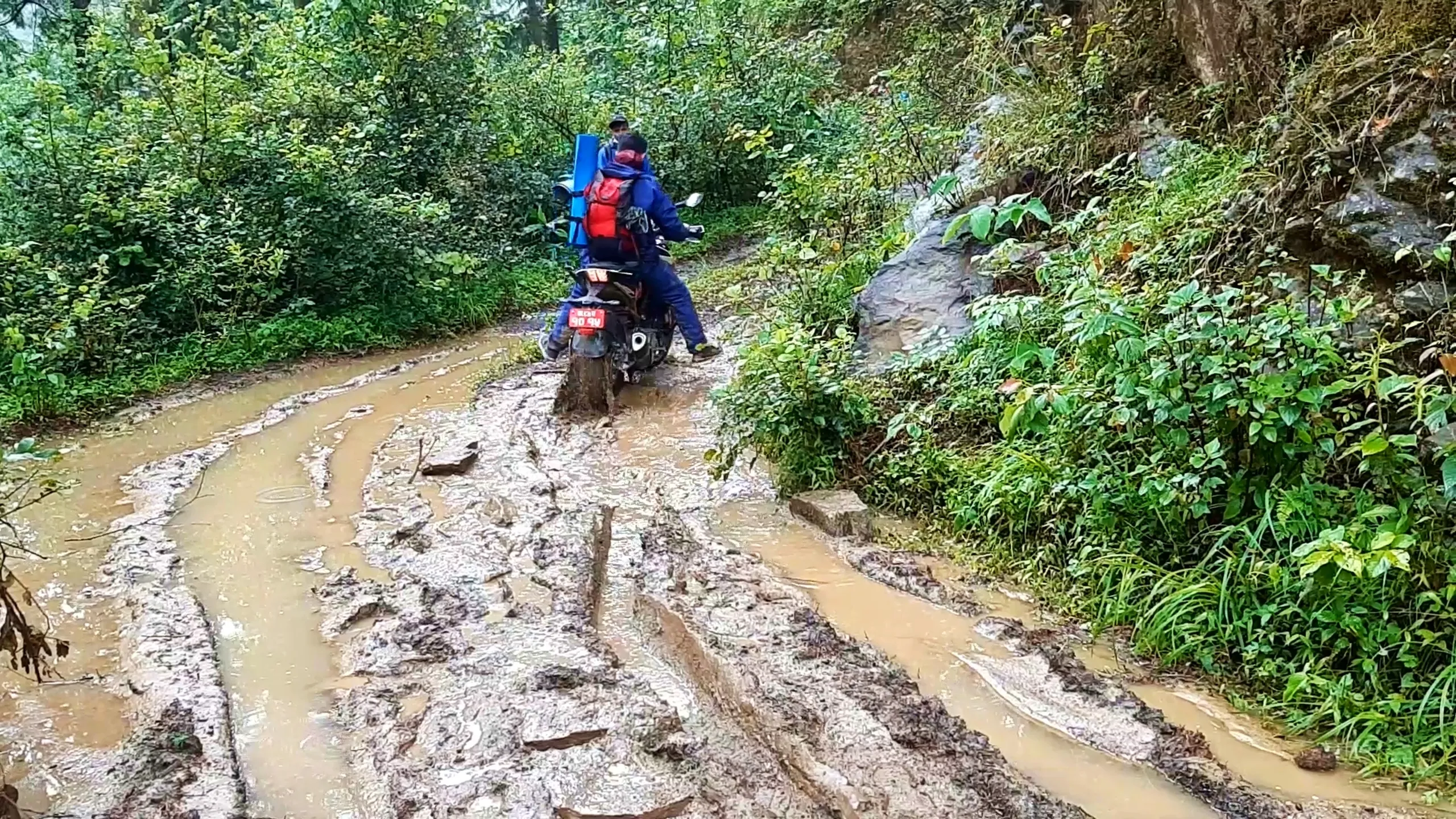 Two friends ride a motorcycle through a flooded, muddy mountain trail in Bethanchowk, Nepal, rear wheel kicking up brown muddy water as it pushes through a deep puddle. The passenger wears a navy blue raincoat with a red-and-black backpack carrying a rolled blue sleeping mat strapped on top, while the rider in dark blue gear leans forward gripping the handlebars. A third hiker in a cap stands ahead on the trail. Lush green forest, ferns, and a rocky hillside frame the narrow dirt path carved with deep tire tracks and rainwater channels. Perfect for Himalayan motorbike trekking, Nepal monsoon adventure, Bethanchowk off-road journey, Kathmandu Valley hiking trail blog content.