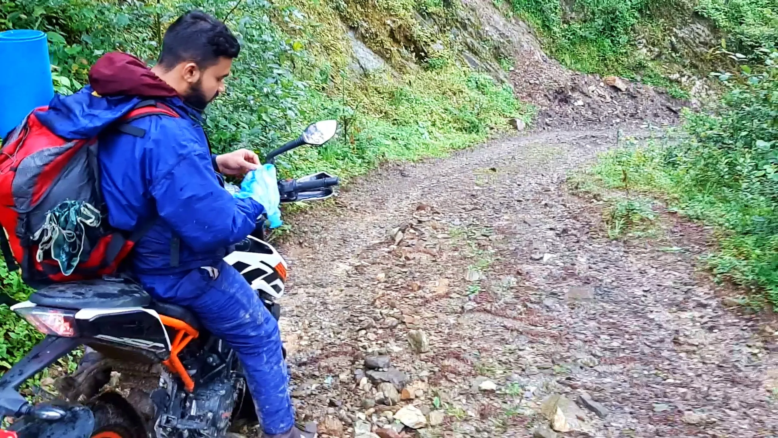 A bearded man in a soaked navy blue raincoat sits sideways on a white and orange motorcycle parked on a gravel mountain trail in Bethanchowk, Nepal, wiping his hands with a light blue cloth. A large red-and-grey trekking backpack with a rolled blue sleeping mat and maroon jacket is strapped to the bike's rear seat. Lush green bushes and a mossy rocky slope rise on the left, while a winding dirt road stretches into the misty forest ahead. Ideal alt text for Himalayan motorbike trekking blog, Nepal off-road adventure ride, Bethanchowk hiking trail, monsoon mountain journey photography.