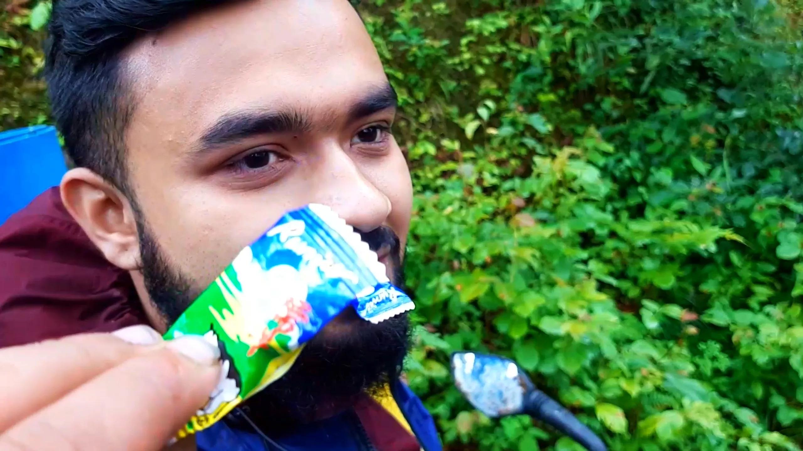 Close-up selfie of a bearded young man with thick dark hair, wearing a maroon hooded jacket over a blue rain jacket, biting open a green, white, and blue foil-wrapped snack or protein bar wrapper with his teeth while smiling, eyes looking off to the side. A blue plastic mat and motorcycle mirror are visible in the blurred background, surrounded by lush green forest foliage. Captures a fun candid moment during a Nepal trekking food break, perfect for Himalayan adventure travel blog, Bethanchowk hiking snacks, monsoon trek lifestyle photography.