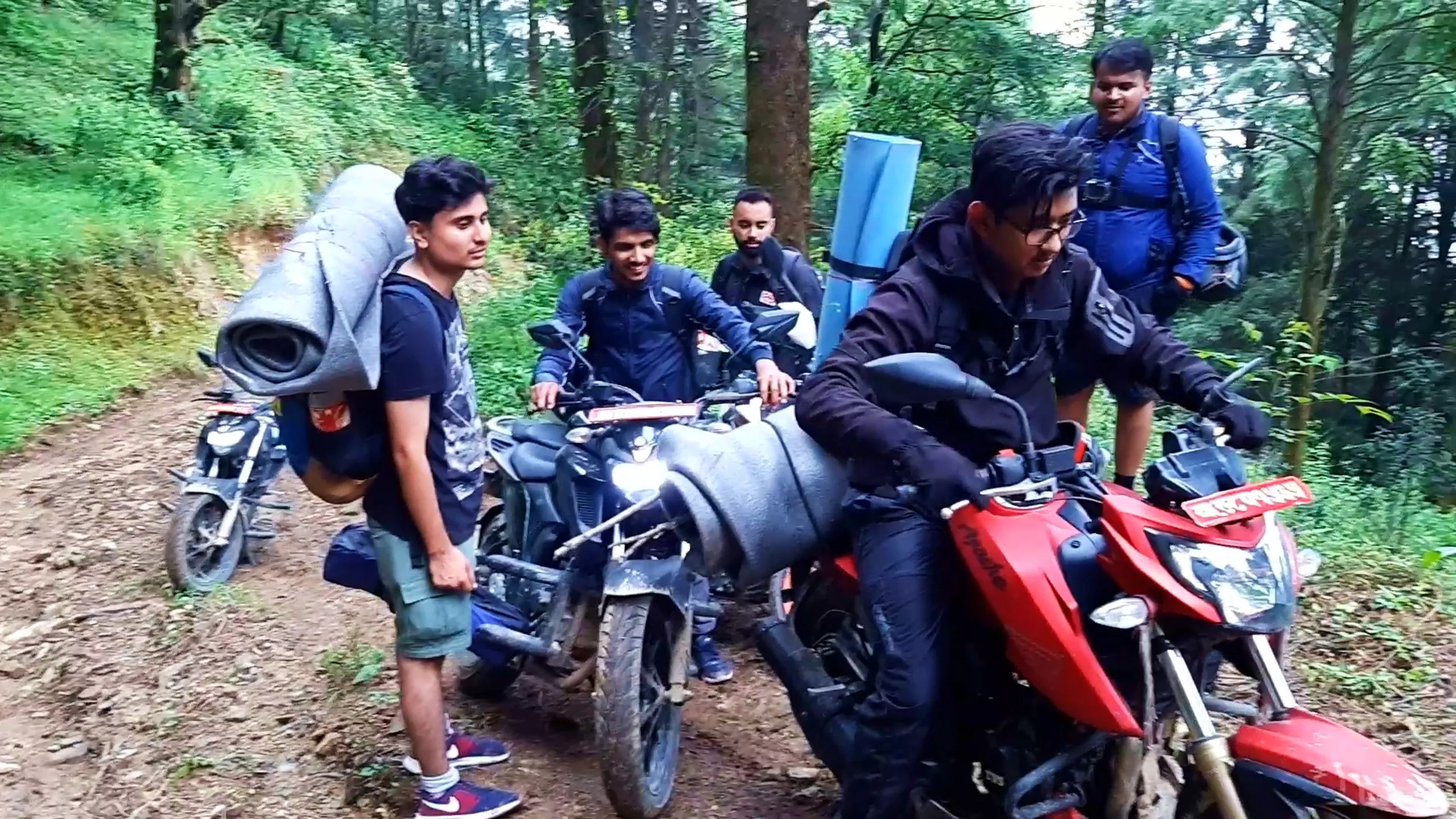 Group photo of five young male friends with their motorbikes parked on a muddy forest trail surrounded by tall green trees during a trekking expedition in Nepal. One man in the front rides a red and black Apache motorbike with a red Nepali number plate, wearing black riding gear and gloves. Behind him, friends stand near other motorbikes, one carrying a large rolled grey sleeping mat strapped to his backpack, another wearing a black jacket with red accents, and a heavier-set man in a blue jacket carrying a large backpack with rolled gear and a motorcycle helmet clipped on. Everyone appears cheerful and relaxed, smiling at the camera amid dense forest greenery and a dirt trail path. This captures a memorable group moment during the Bethanchowk motorbike and hiking trip, friends adventure travel Nepal, forest trail biking Kathmandu Valley, group trekking expedition Himalayas.