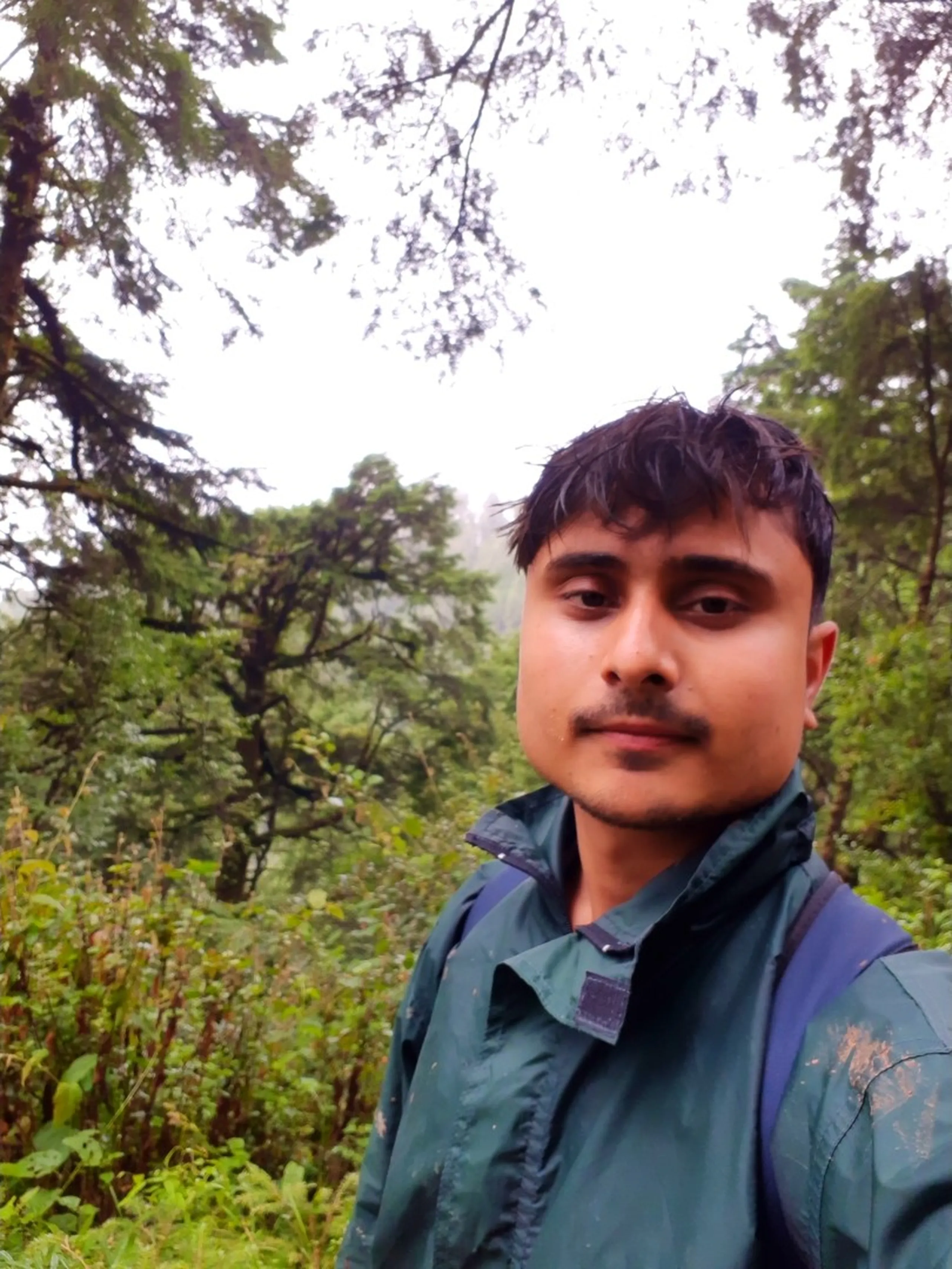 A close-up selfie of a young man with tousled dark hair and a light mustache, wearing a dark green rain jacket with a blue backpack strap visible over his shoulder, looking calmly at the camera. Behind him, tall pine and fir trees rise into a foggy white sky, with dense green forest foliage surrounding the misty Himalayan trail in Bethanchowk, Nepal. A candid trekking portrait ideal for Nepal Himalayan adventure blog, Bethanchowk hiking journey, monsoon trek selfie photography, Kathmandu Valley forest trail content.