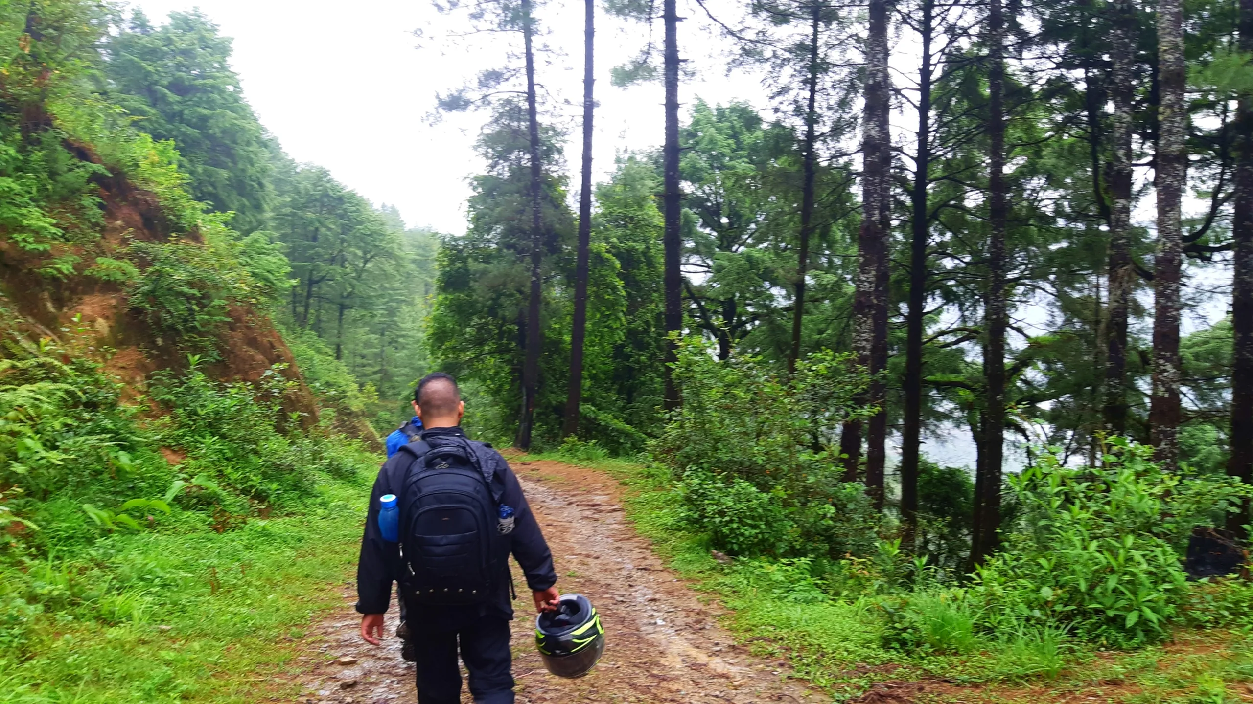 A hiker in a black rain jacket with a black trekking backpack carrying two blue water bottles walks along a reddish dirt forest trail in Bethanchowk, Nepal, holding a black-and-neon-green motorcycle helmet in his hand. Ahead, another hiker in a blue jacket is partially visible on the path. Tall pine trees line both sides of the winding trail, with a misty grey-green valley and hazy mountains visible through the trees under an overcast monsoon sky. Great for Nepal Himalayan trekking blog, Bethanchowk forest hike, pine woodland trail photography, Kathmandu Valley adventure trek content.