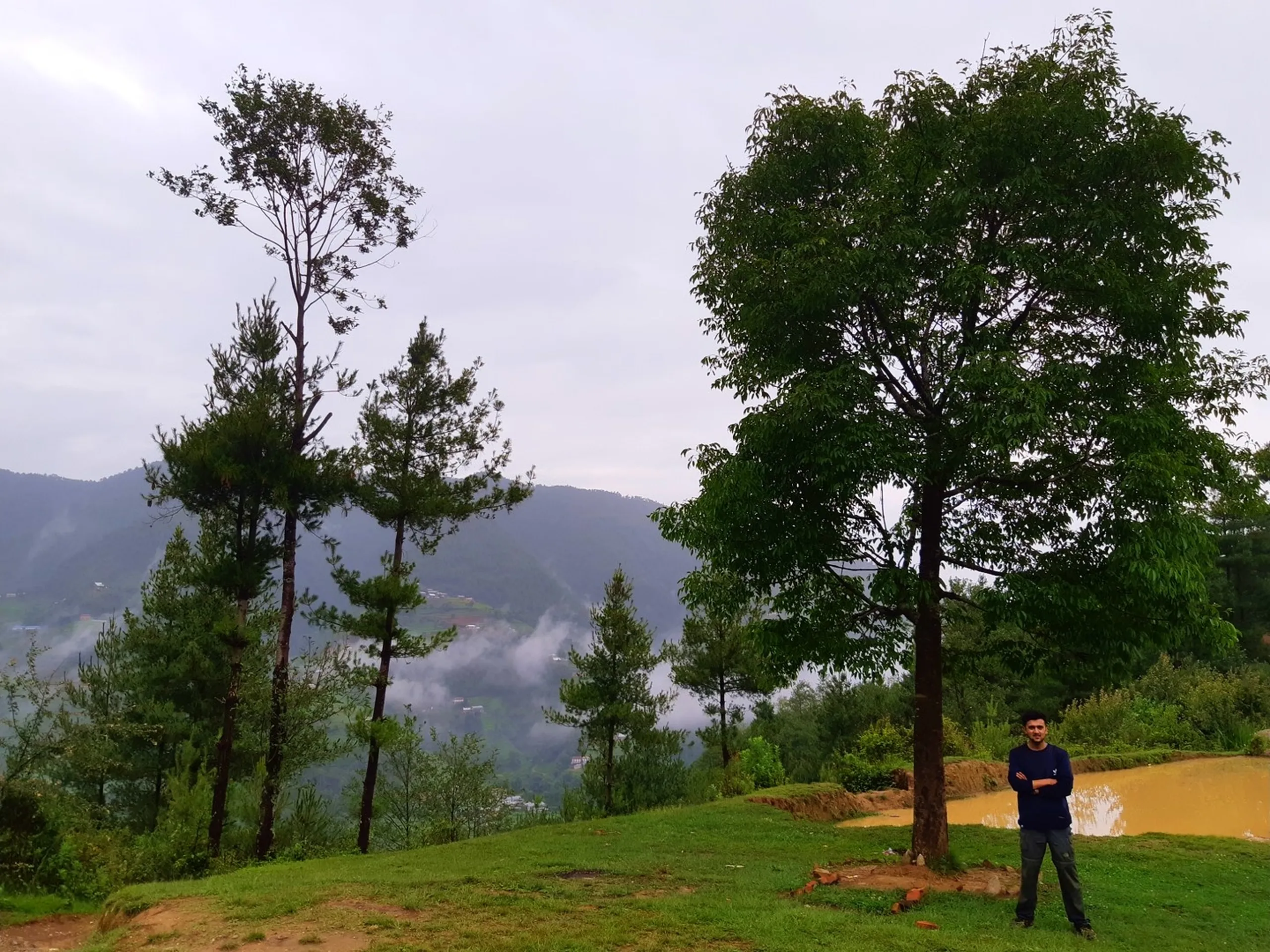 A young man in a dark blue sweater and grey trousers stands with arms crossed beside a tall leafy tree on a grassy hilltop in Bethanchowk, Nepal, smiling at the camera. Beside him, a muddy orange water pond sits at the edge of the clearing. In the background, several slender pine trees stand against a vast misty mountain landscape, with layered hills, fog, and a distant valley village fading into the cloudy sky. Ideal for Nepal Himalayan trekking blog, Bethanchowk viewpoint travel photography, monsoon mountain scenery, Kathmandu Valley adventure content.