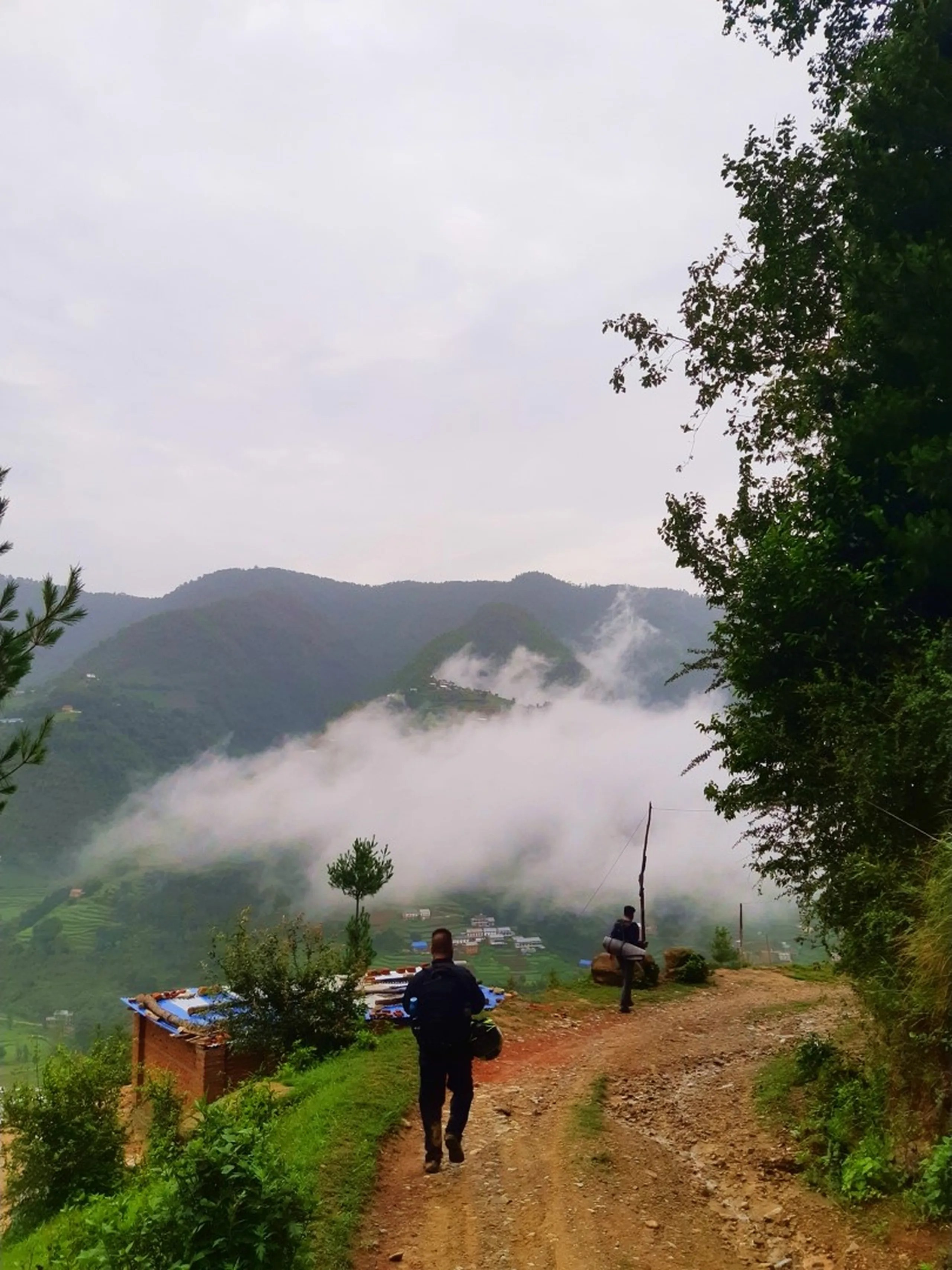 A hiker in a black jacket carrying a dark backpack with a green item strapped on top walks down a reddish dirt path on a misty hillside in Bethanchowk, Nepal, heading toward a small roadside shop with a blue tarp roof. Below, thick white clouds blanket a deep valley, partially revealing terraced green hillsides and a scattered village with colorful rooftops. Another person stands further down the slope near a wooden pole. Lush trees frame the scene under an overcast monsoon sky. Perfect for Nepal Himalayan trekking blog, Bethanchowk village viewpoint, misty mountain landscape photography, Kathmandu Valley hiking adventure content.