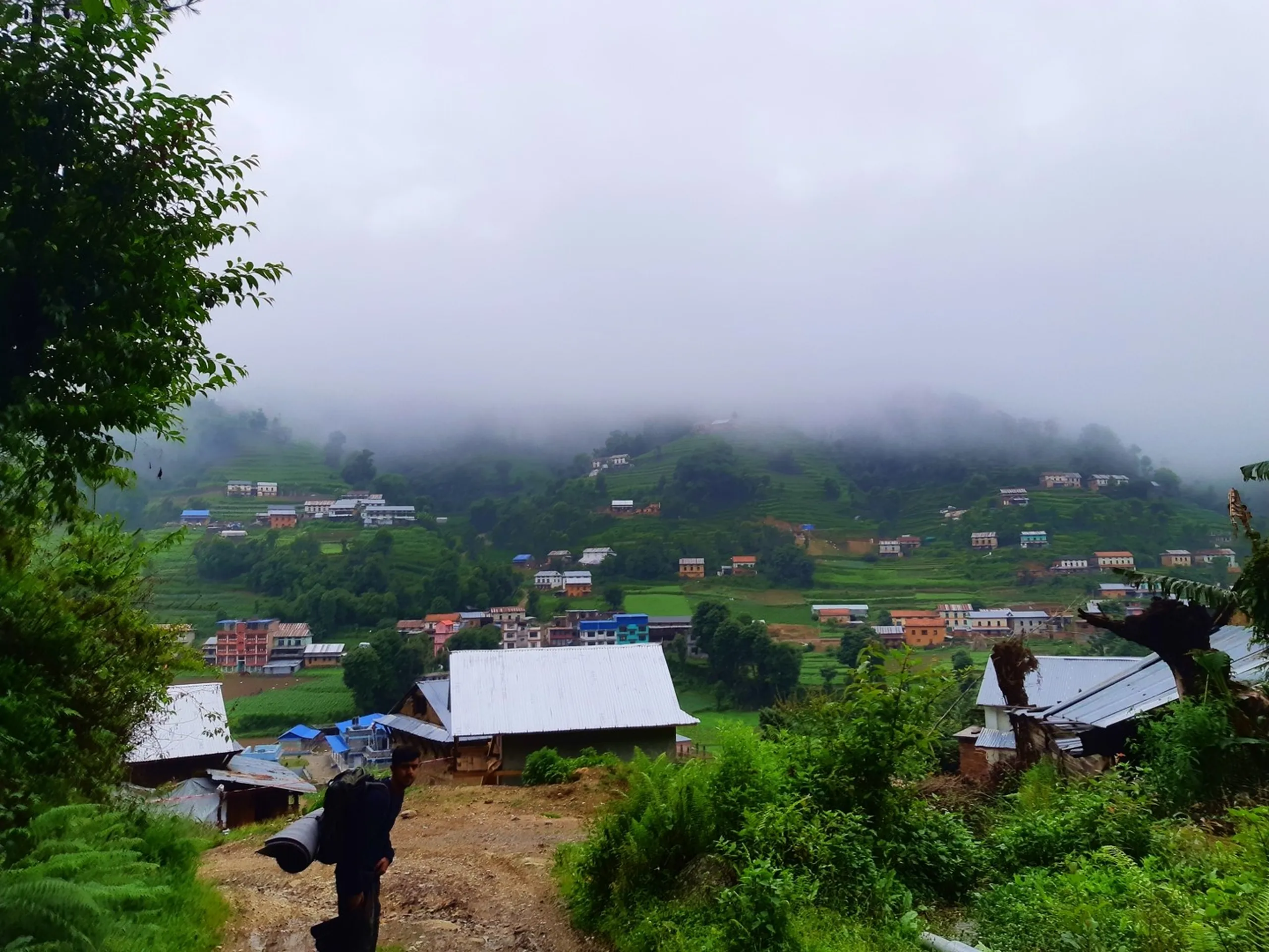 A hiker carrying a large dark backpack with a rolled mat strapped on top walks down a dirt path toward a sprawling terraced village in a misty Himalayan valley near Bethanchowk, Nepal. Below, dozens of colorful houses with blue, white, and red tin roofs dot lush green terraced farmland and rice fields, with thick clouds rolling over the forested hills above the settlement. Dense green ferns and trees frame the foreground on both sides of the trail. A breathtaking wide-angle shot perfect for Nepal Himalayan village trekking blog, Bethanchowk hiking trail, monsoon mountain valley photography, Kathmandu Valley rural landscape content.