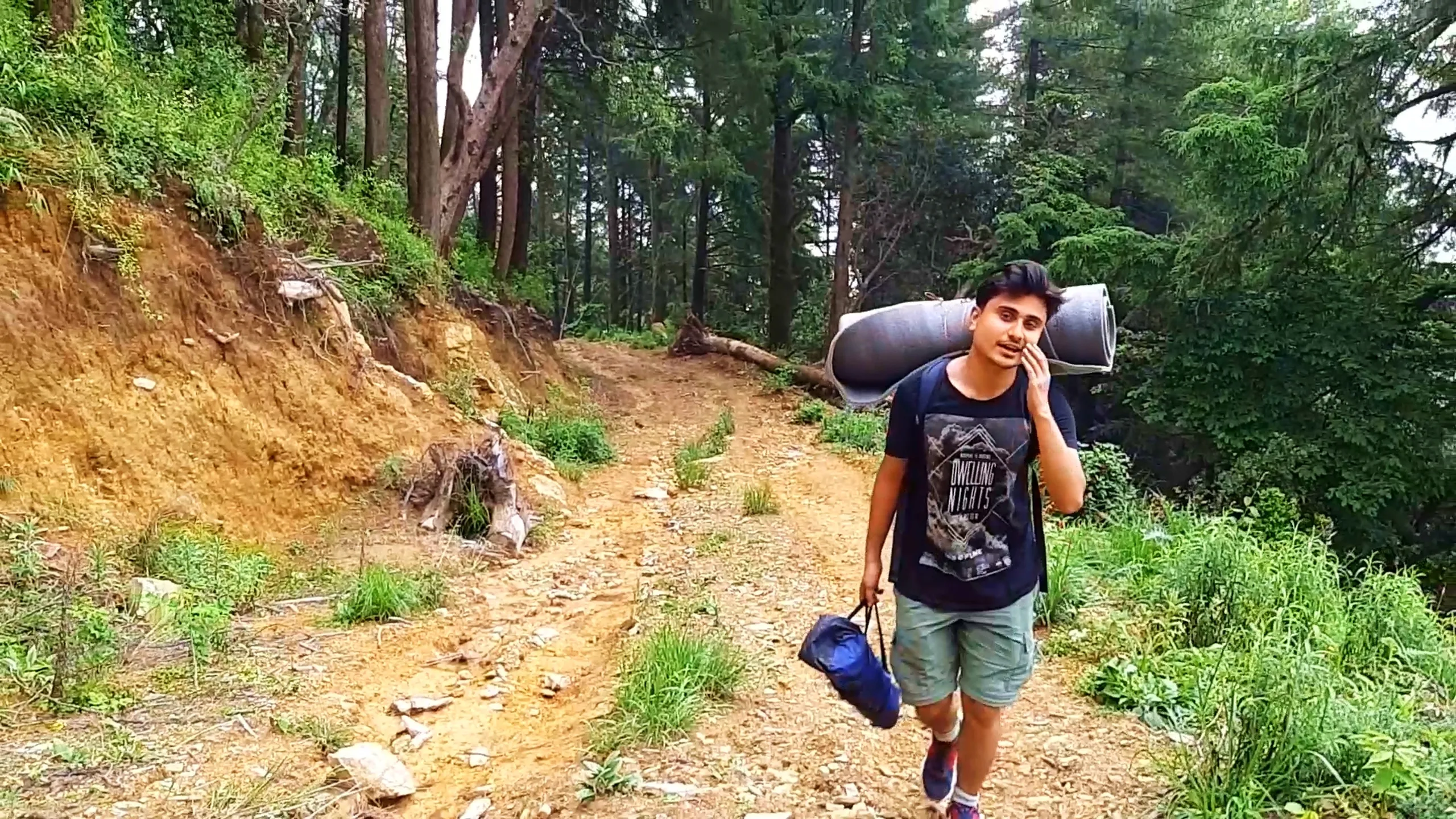 Photo of a young man walking along a forest dirt trail carrying a large rolled grey sleeping mat strapped horizontally across his backpack and a blue drawstring bag in his hand, wearing a black "Dwelling Nights" graphic t-shirt, olive green cargo shorts, and red sneakers, touching his face with one hand while talking. The trail winds through tall pine and cedar trees with a steep eroded earthen slope on the left covered in green undergrowth, and dense forest vegetation on the right. This image shows a hiker trekking through pine forest trails near Bethanchowk, camping gear backpacking Nepal, forest hiking trail Kathmandu hills, Himalayan jungle trekking adventure.