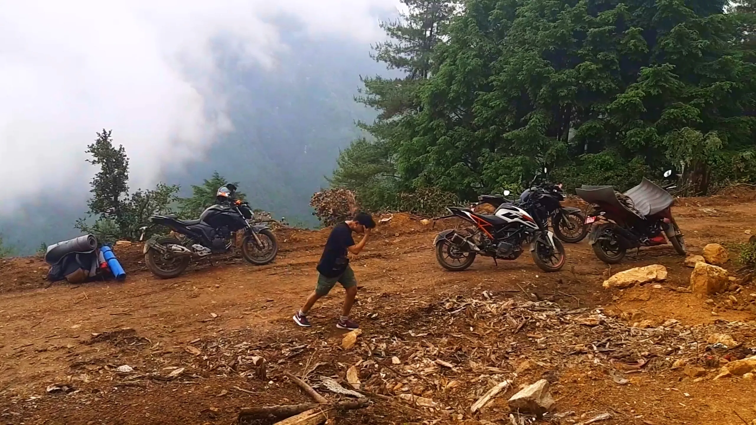 Wide landscape photo of three motorbikes parked on a dirt clearing at the edge of a misty mountain ridge in Bethanchowk, Nepal, with thick white fog and clouds rolling over the valley below on the left side. The motorbikes include a black naked sportbike with a helmet resting on its seat, a white and orange KTM Duke style bike, and another bike with cargo bags strapped to the back. A young man wearing a black t-shirt, olive green shorts, and sneakers walks across the dirt ground with his hand covering his face, near a pile of camping gear including rolled sleeping mats and backpacks on the left. Dense pine trees line the right side of the ridge. This captures the dramatic cloud-covered mountain camping spot at Bethanchowk highest point, foggy Himalayan viewpoint Nepal, motorbike camping trip Kathmandu Valley, misty mountain trekking destination.