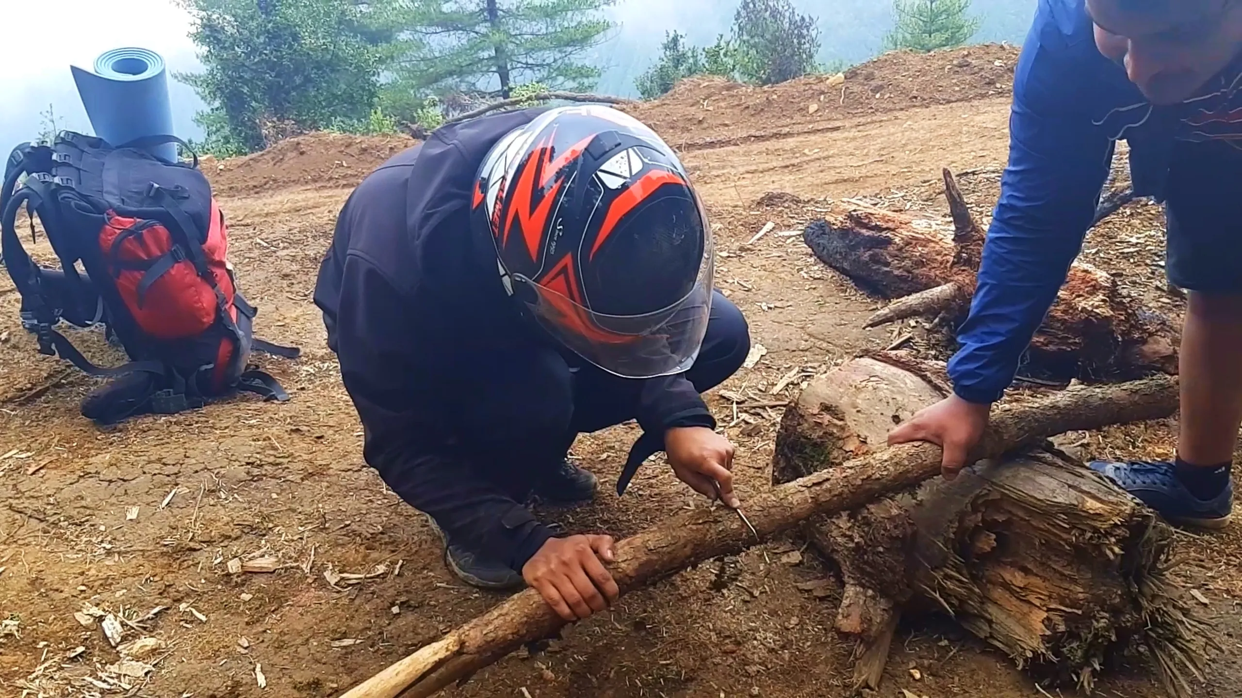 Close-up photo of a man wearing a black and orange graphic full-face motorcycle helmet with a clear visor and a navy blue riding jacket, crouching down to break or saw a wooden log for firewood using a small tool, while another person in a blue jacket helps hold the log steady on the dirt ground. Beside them lies a red backpack with a light blue rolled sleeping mat strapped on top. The misty forested mountain background is visible through fog, with green pine trees on the upper right. This shows campers collecting firewood for a bonfire during Bethanchowk overnight camping trip, Nepal jungle camping preparation, Himalayan campfire wood gathering, adventure camping Kathmandu hills.