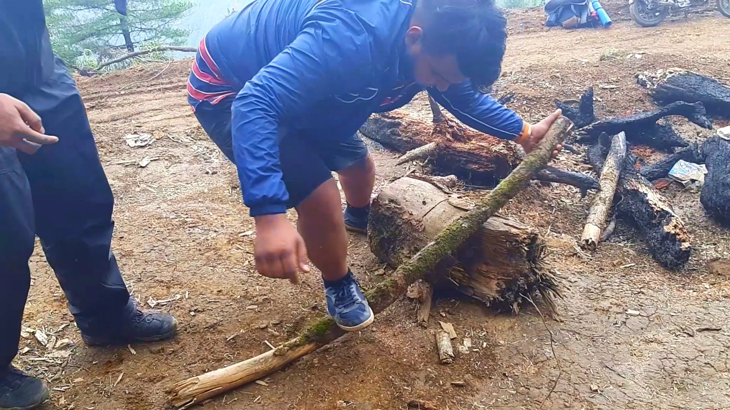 Photo of a man wearing a blue jacket with red and white striped detailing and blue sneakers, bending over to break a thick mossy wooden log over a tree stump near a burnt campfire pit filled with charred black wood logs and ash. Another person stands to the left wearing dark pants and boots, partially visible, gesturing with their hand. In the background, fog-covered pine trees and a glimpse of a parked motorbike with a sleeping mat are visible on a dirt clearing. This depicts firewood preparation for an overnight bonfire camp at Bethanchowk highest point, Nepal camping survival skills, Himalayan jungle camp setup, group camping trip Kathmandu hills.