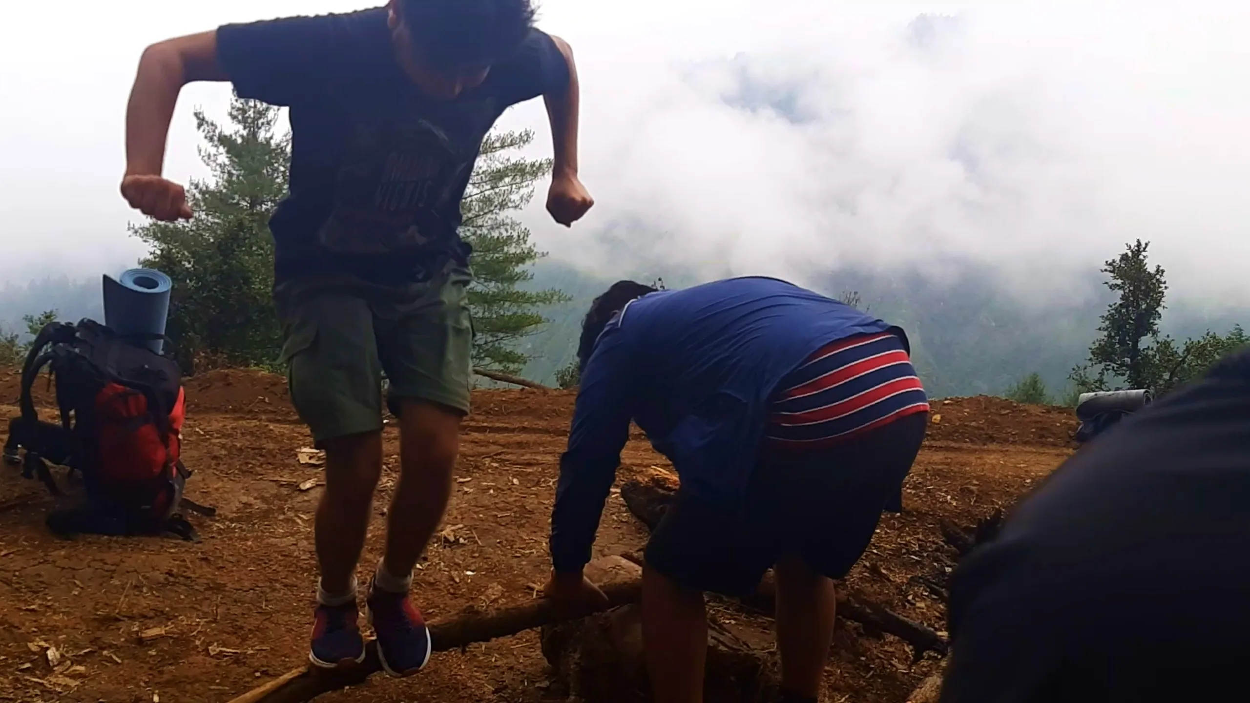 Dynamic action shot of a young man in a navy blue graphic t-shirt and olive green cargo shorts jumping mid-air with arms raised in excitement on a dirt clearing, while another man wearing a blue jacket with red and white stripes bends over working on a wooden log near a burnt campfire pit. A red backpack with a light blue rolled sleeping mat sits on the ground to the left, and another person's dark sleeve is visible in the foreground. Thick white fog and mist fill the background completely obscuring any mountain view. This captures playful high spirits during firewood collection at Bethanchowk camping spot, fun moments Nepal trekking trip, foggy mountain camp Kathmandu Valley, adventure camping memories Himalayas.