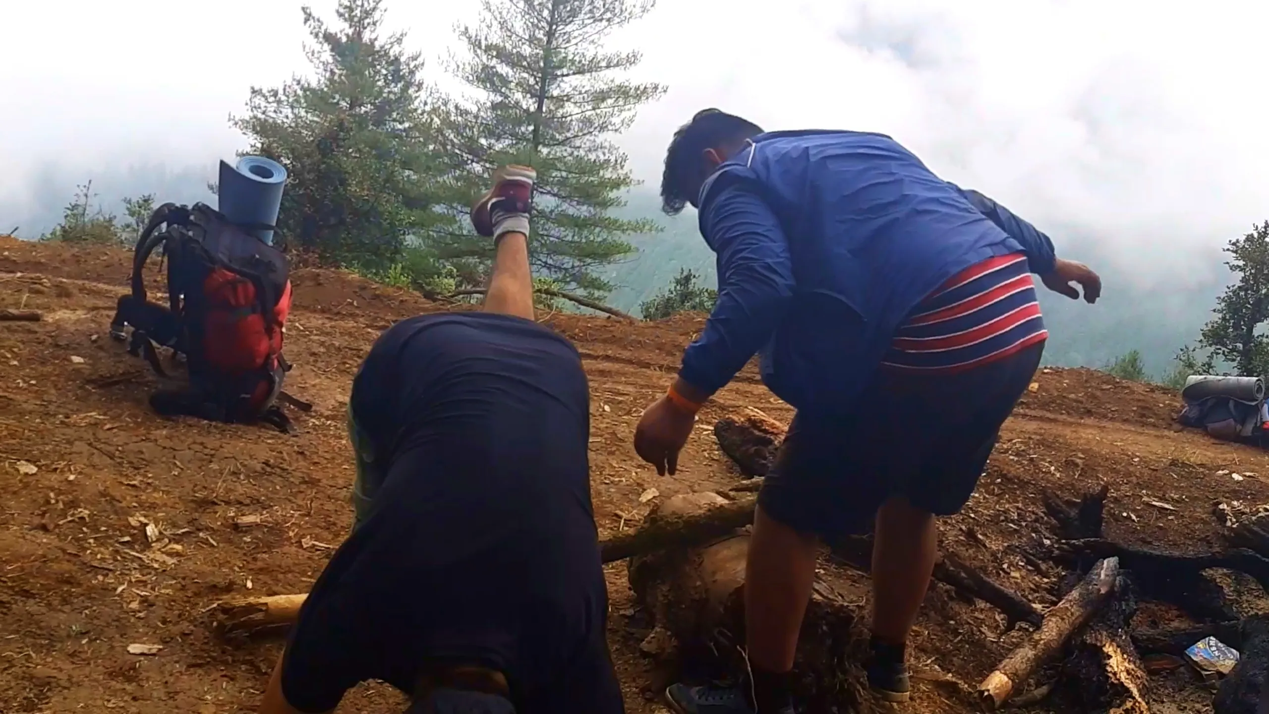 Action photo taken from a low angle showing a man's leg kicking up mid-motion wearing a maroon sneaker, while another man in a blue and red striped jacket bends down working with wooden logs near a campfire pit with burnt charred wood. A red backpack with a rolled light blue sleeping mat sits on the dirt ground to the left, with a small green pine tree and thick white fog covering the misty mountain background. Another rolled sleeping mat and motorbike parts are visible on the right edge of the frame. This shows energetic camp chores and firewood gathering in foggy conditions at Bethanchowk overnight stop, Nepal adventure camping fun, Himalayan trekking group activities, misty mountain campsite Kathmandu hills.