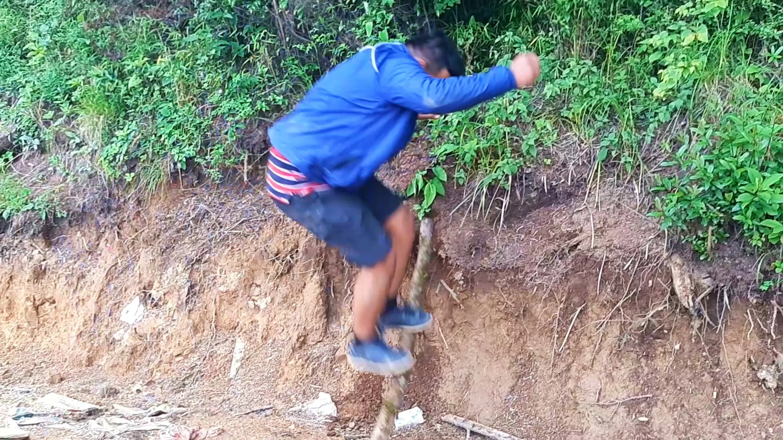 Action shot of a young man in a blue jacket with red and white striped shorts jumping or balancing on a thick wooden log positioned over an eroded dirt slope, with one arm raised forward in motion and wearing blue sneakers. The background shows a steep reddish-brown eroded earthen embankment covered with dense green shrubs, ferns, and wild vegetation typical of Himalayan forest terrain. Scattered litter and dirt clods are visible on the ground. This captures playful balancing antics on fallen logs during Bethanchowk forest trekking, Nepal jungle adventure games, off-trail hiking fun Kathmandu hills, Himalayan forest exploration.