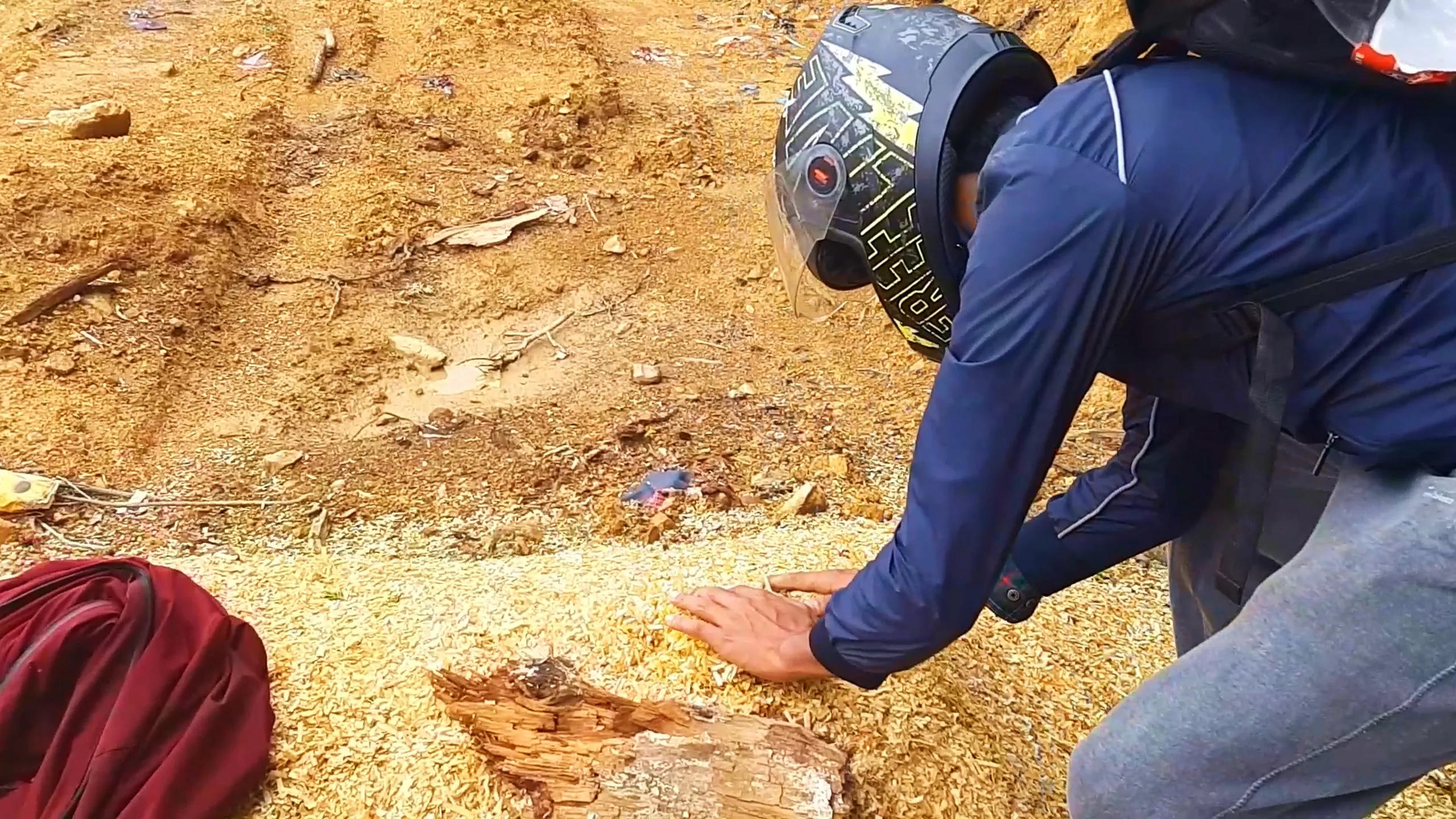Close-up shot of a man wearing a black and yellow graphic full-face motorcycle helmet with a clear visor flipped up and a navy blue riding jacket with a grey backpack, crouching down and reaching his hand out to touch a pile of golden-brown wood shavings or sawdust scattered across the ground near a cut tree stump. A maroon red backpack is visible in the bottom left corner. The dirt ground is covered in scattered wood debris, dry leaves, and small twigs. This shows close inspection of fresh-cut firewood at Bethanchowk camping site, Nepal trekking campfire preparation, sustainable wood collection Himalayan camping, adventure trip details Kathmandu hills.