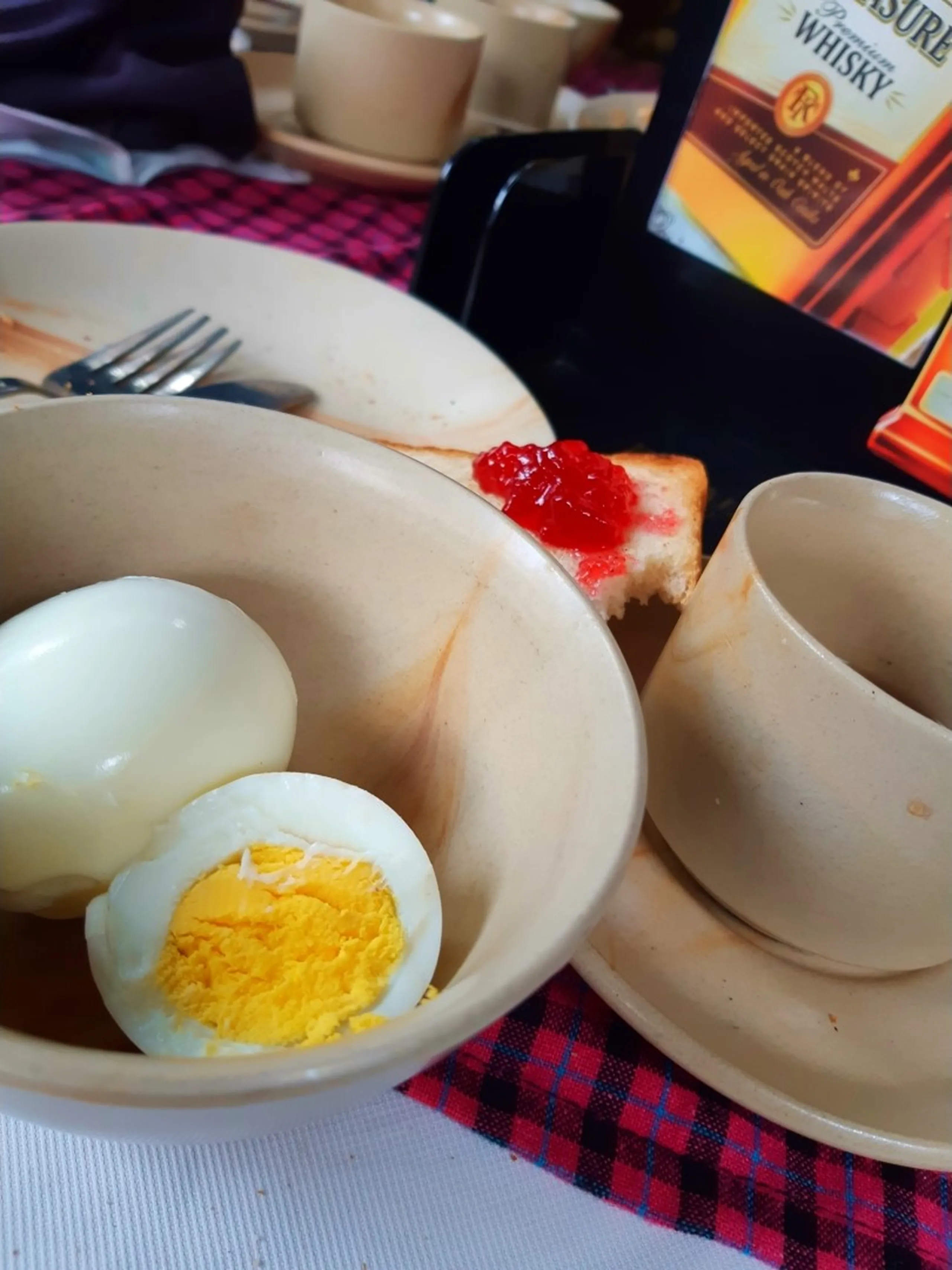 Close-up shot of a rustic breakfast table setting during a Nepal trekking road trip, featuring a ceramic bowl with two peeled hard-boiled eggs cut in half showing bright yellow yolks, a slice of white bread toast spread with bright red strawberry jam on a black plate, an empty beige ceramic teacup on a matching saucer, and a stainless steel fork resting on a plate edge. The table is covered with a red and black checkered cloth napkin and white tablecloth. In the background blurred, a bottle of 8PM Premium Whisky stands beside another stacked teacup and saucer. This image represents a typical Nepali breakfast spread enjoyed during a motorbike hiking trip to Bethanchowk, Himalayan trekking food culture, Nepal travel breakfast, road trip snacks Nepal.