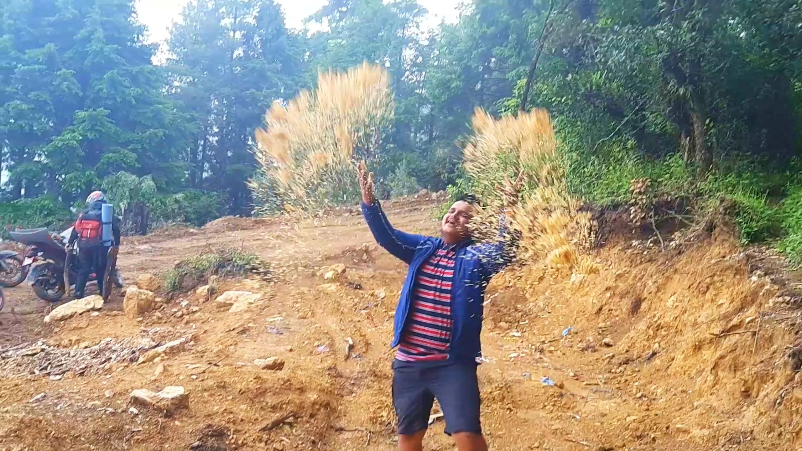 Playful action shot of a young man in a blue zip-up jacket with red and navy striped t-shirt and dark shorts joyfully throwing handfuls of golden hay, dried grass, or wood shavings into the air with both arms raised, creating a dramatic spray of golden particles against a backdrop of dense green pine forest. In the background, another hiker wearing a helmet and red backpack with a blue rolled sleeping mat stands near a parked motorbike on a muddy dirt clearing. Misty sunlight filters through the trees creating a glowing atmospheric effect. This captures a joyful celebratory moment during Bethanchowk forest camping trip, Nepal adventure travel happiness, Himalayan trekking fun activities, group hiking high spirits Kathmandu hills.