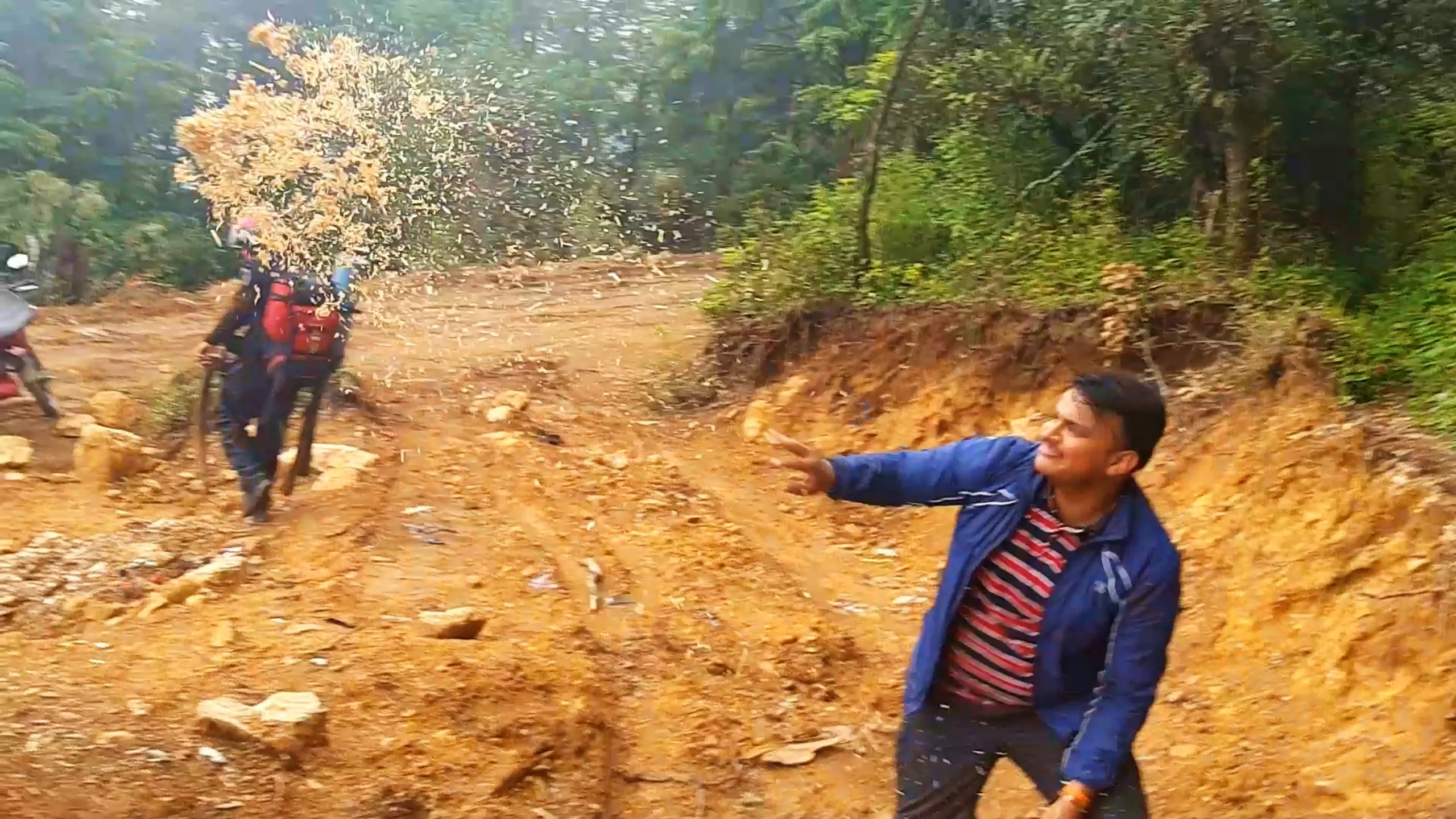 Dynamic action photo of a young man in a blue jacket with red and navy striped t-shirt mid-throw, flinging a spray of golden wood shavings or hay into the air with his arm extended, smiling widely on a muddy orange dirt clearing surrounded by dense green forest. In the background, another hiker wearing a black helmet, red backpack, and blue rolled sleeping mat walks near a parked motorbike, partially obscured by the flying debris cloud. The eroded reddish dirt embankment and green bushes frame the scene. This captures spontaneous fun and camping preparation excitement at Bethanchowk highest point, Nepal jungle trekking adventure, Himalayan camping group activities, motorbike trekking memories Kathmandu hills.