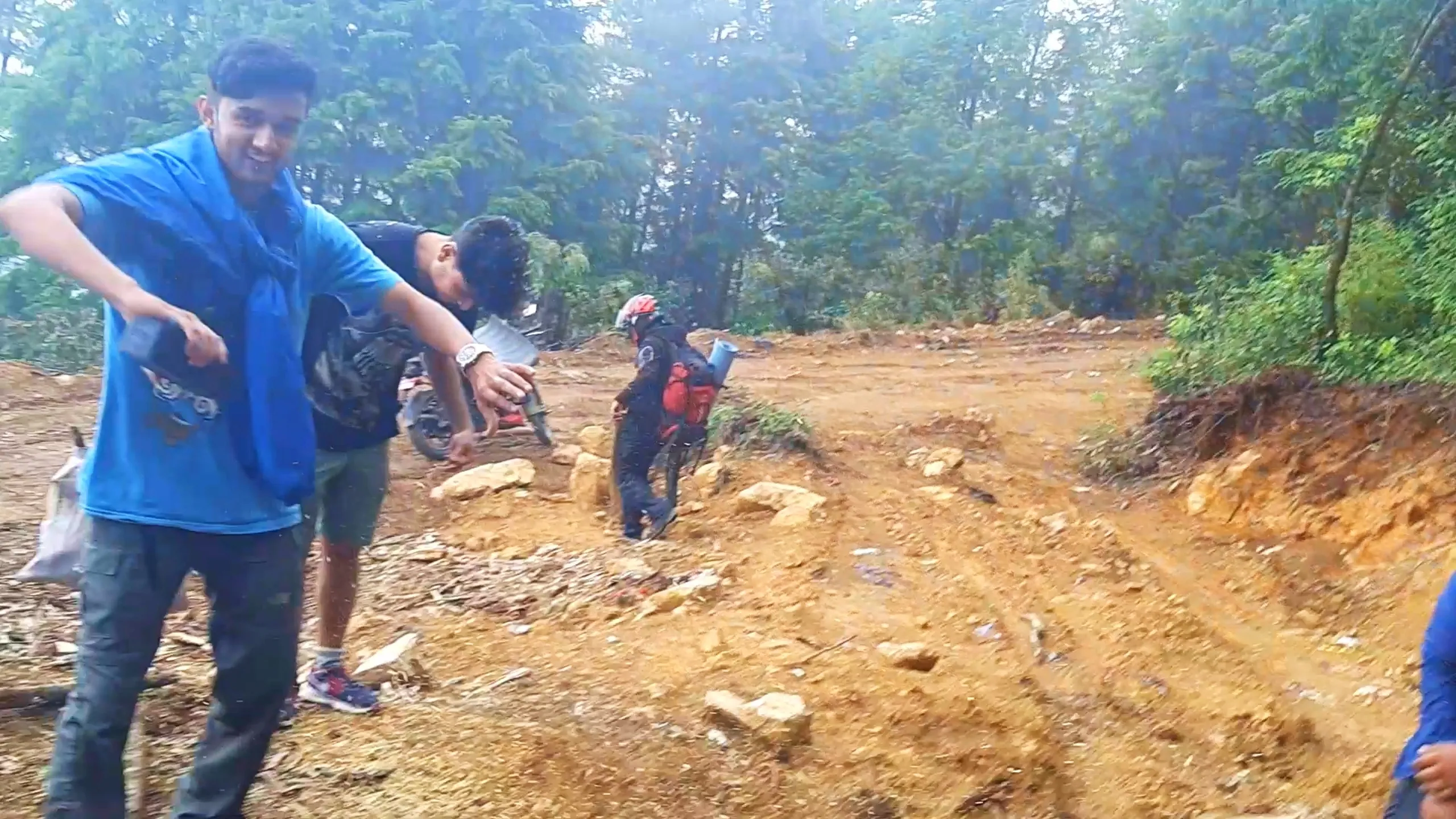 Candid group photo of three young male friends on a misty forest dirt trail during a Nepal trekking trip, captured in slightly blurred motion. The man in front wears a bright blue t-shirt with a graphic print and grey cargo pants, smiling and gesturing with one hand while holding a dark object. Behind him, another friend in a black graphic t-shirt and green shorts bends down near a parked motorbike, while a third hiker wearing a black and red riding jacket with a black helmet, red backpack, and rolled sleeping mat walks toward the dense green forest. The ground is covered in reddish-orange dirt with scattered rocks and forest debris. Misty pine trees fill the background under hazy daylight. This depicts a candid group moment during Bethanchowk forest trekking trip, Nepal friends adventure travel, Himalayan jungle hiking group, motorbike camping expedition Kathmandu hills.