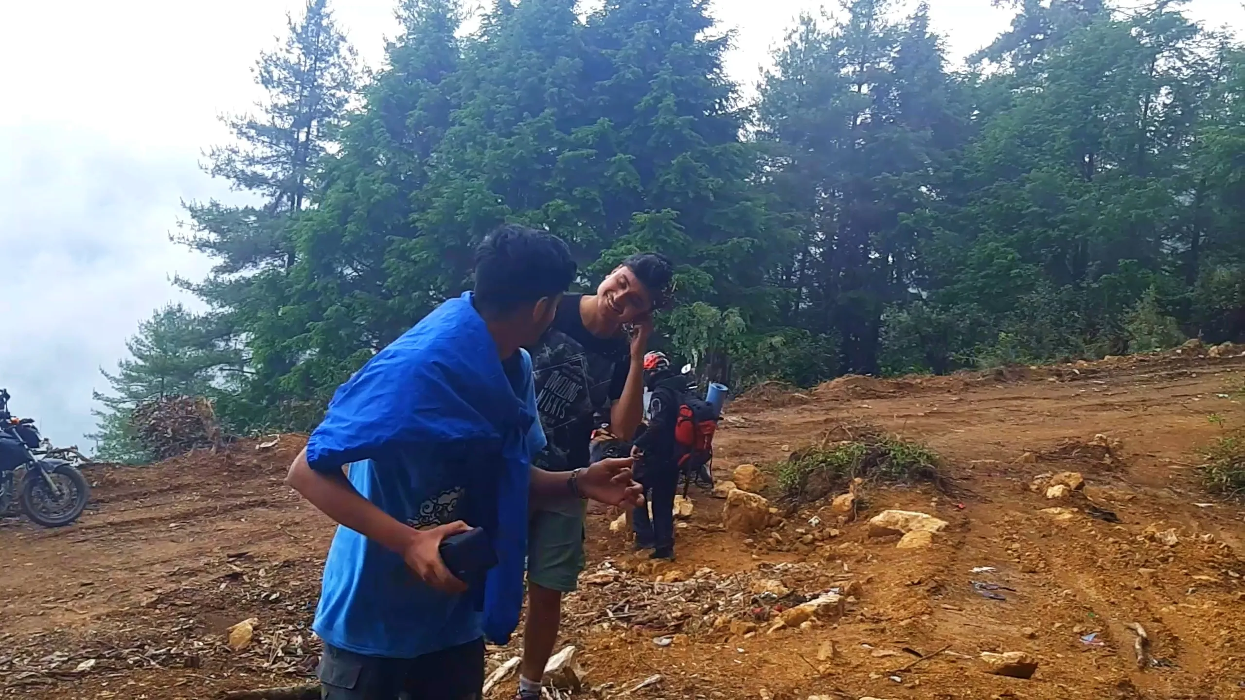Warm candid photo of two young men sharing a laugh on a misty mountain dirt clearing surrounded by tall pine and cedar trees. The man in front wears a blue t-shirt draped loosely over his shoulders like a cape with cargo shorts, holding a dark object in one hand, while his friend beside him in a black "Dwelling Nights" graphic t-shirt smiles and touches his face playfully. In the background, a third hiker wearing a black helmet and red backpack with rolled sleeping mat walks near a parked motorbike, with another motorbike visible on the left edge. Thick fog and mist roll through the dense pine forest backdrop. This captures friendly bonding moments during Bethanchowk camping trip, Nepal trekking friendship memories, Himalayan misty forest adventure, group hiking laughter Kathmandu hills.