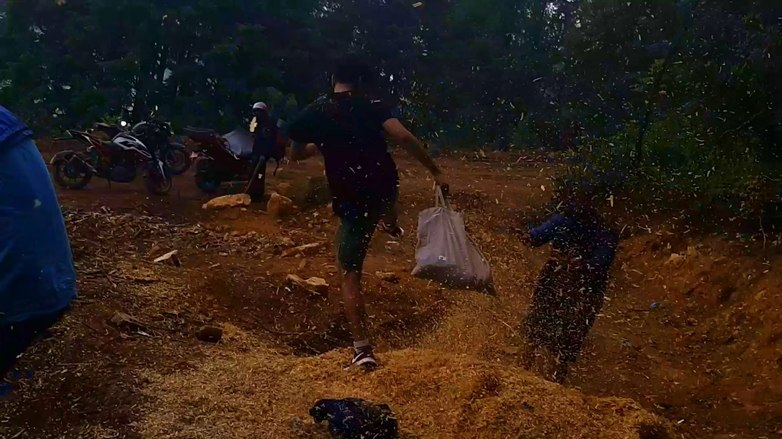 Blurry dark action shot of a young man in a black t-shirt and dark green shorts running across a dirt clearing covered in scattered golden wood shavings and debris flying through the air, carrying a white plastic bag in one hand during dim evening or low-light forest conditions. In the background, two motorbikes are parked near a person wearing a white helmet, with another figure partially visible on the right side amid the flying debris cloud. A blue rain jacket is visible in the foreground corner. Dense dark forest trees fill the background under fading daylight. This captures energetic evening camp activity at Bethanchowk forest campsite, Nepal trekking dusk adventure, Himalayan camping preparation chaos, motorbike trip group dynamics Kathmandu hills.