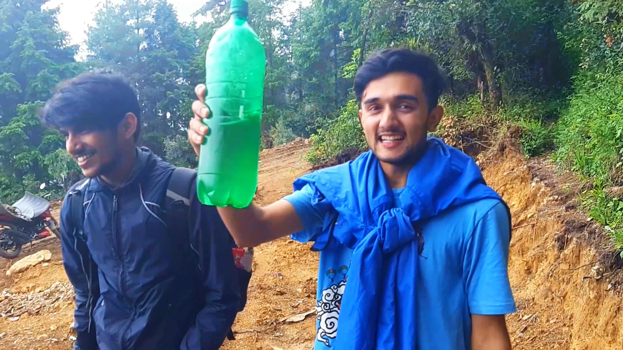 Cheerful close-up photo of two young men standing on a misty forest dirt trail, with one man on the right wearing a sky-blue t-shirt with a rain jacket draped around his shoulders, smiling broadly while holding up a green plastic bottle filled with liquid. Beside him, another friend with short hair wears a navy blue jacket with a backpack, also smiling. Dense green pine and cedar trees fill the misty background with a parked motorbike visible on the lower left edge of the frame. Soft diffused sunlight creates a hazy glow through the fog. This captures a joyful refreshment break during Bethanchowk forest trekking trip, Nepal hiking friends camaraderie, Himalayan misty mountain adventure, group camping break Kathmandu hills.