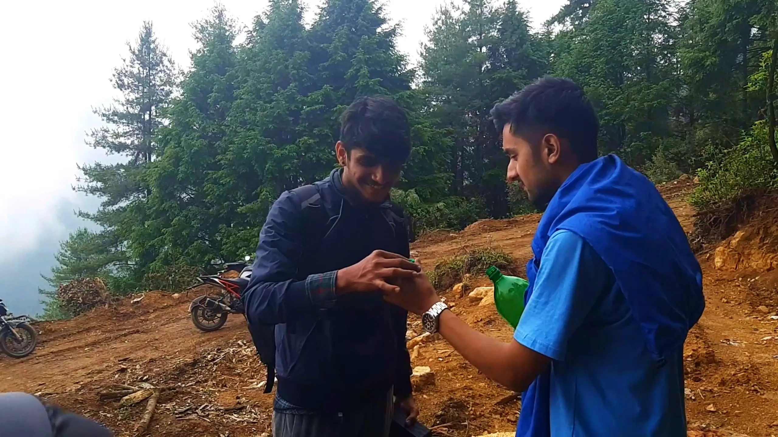 Close-up candid photo of two young men standing together on a misty mountain dirt trail surrounded by tall pine and cedar trees. The man on the left wears a dark navy jacket with a plaid shirt collar visible and a backpack, smiling and looking down at his hands. Beside him, a friend in a sky-blue t-shirt with a blue rain jacket draped around his shoulders holds a green plastic bottle and a silver wristwatch, gesturing toward the other man's hands. A parked motorbike with KTM Duke styling is visible in the background on the dirt path, with dense fog-covered pine forest filling the misty backdrop. This captures a friendly interaction moment during Bethanchowk forest trekking trip, Nepal hiking buddies adventure, Himalayan misty mountain camping, group bonding Kathmandu hills.