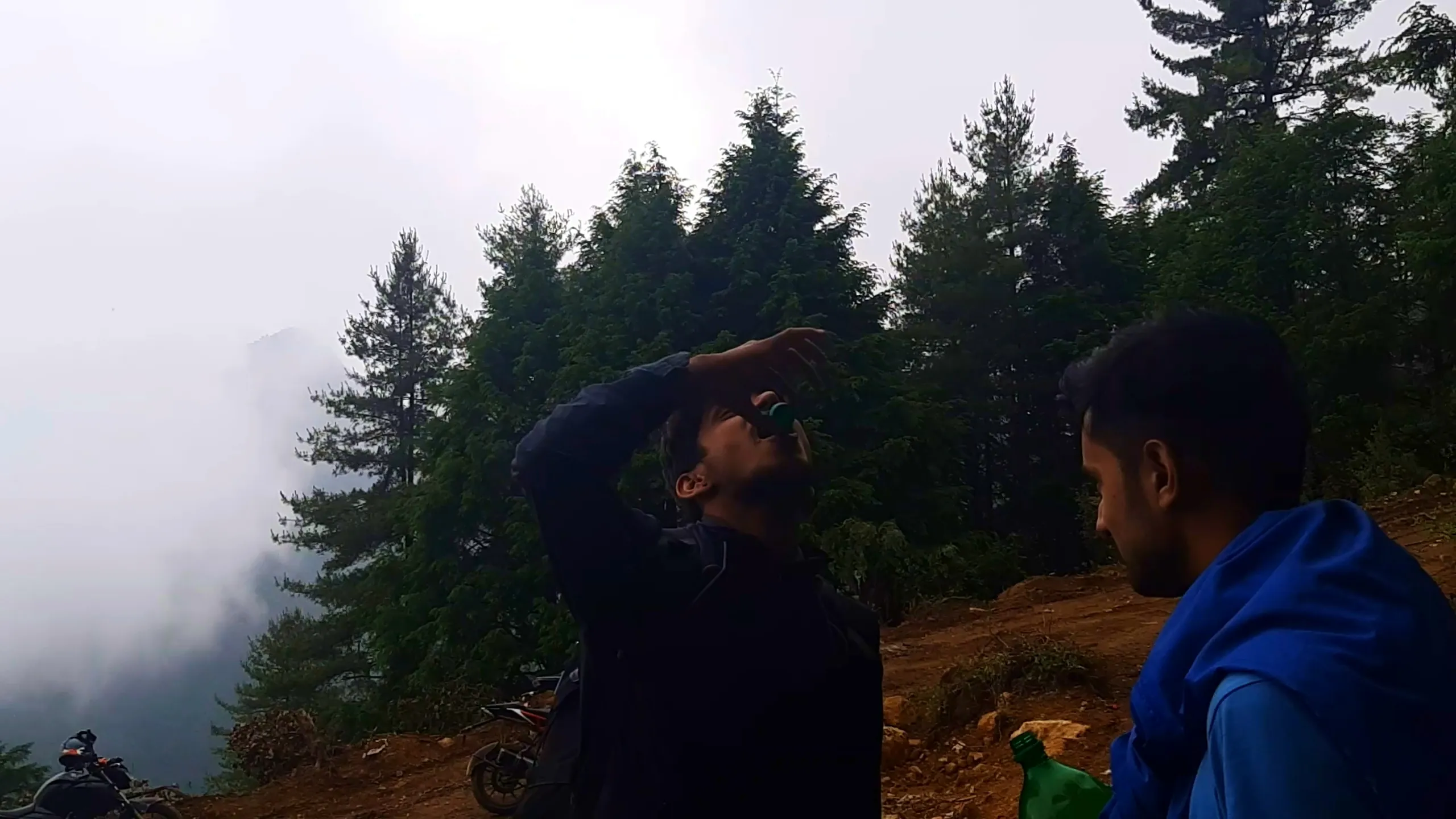 Atmospheric photo of two young men standing on a misty mountain ridge surrounded by tall pine and cedar trees shrouded in thick fog. The man in the center wears a dark navy jacket, tilting his head back with one arm raised to drink from a small green bottle, his silhouette darkened against the bright foggy sky. Beside him on the right, another man in a blue rain jacket sits or stands holding a green plastic bottle, looking towards his friend. Parked motorbikes are visible on the dirt ground in the lower left of the frame, with dense pine forest fading into white mist in the background. This captures a quiet refreshment moment amid heavy fog at Bethanchowk highest point, Nepal misty mountain trekking, Himalayan adventure camping break, foggy forest hiking Kathmandu hills.