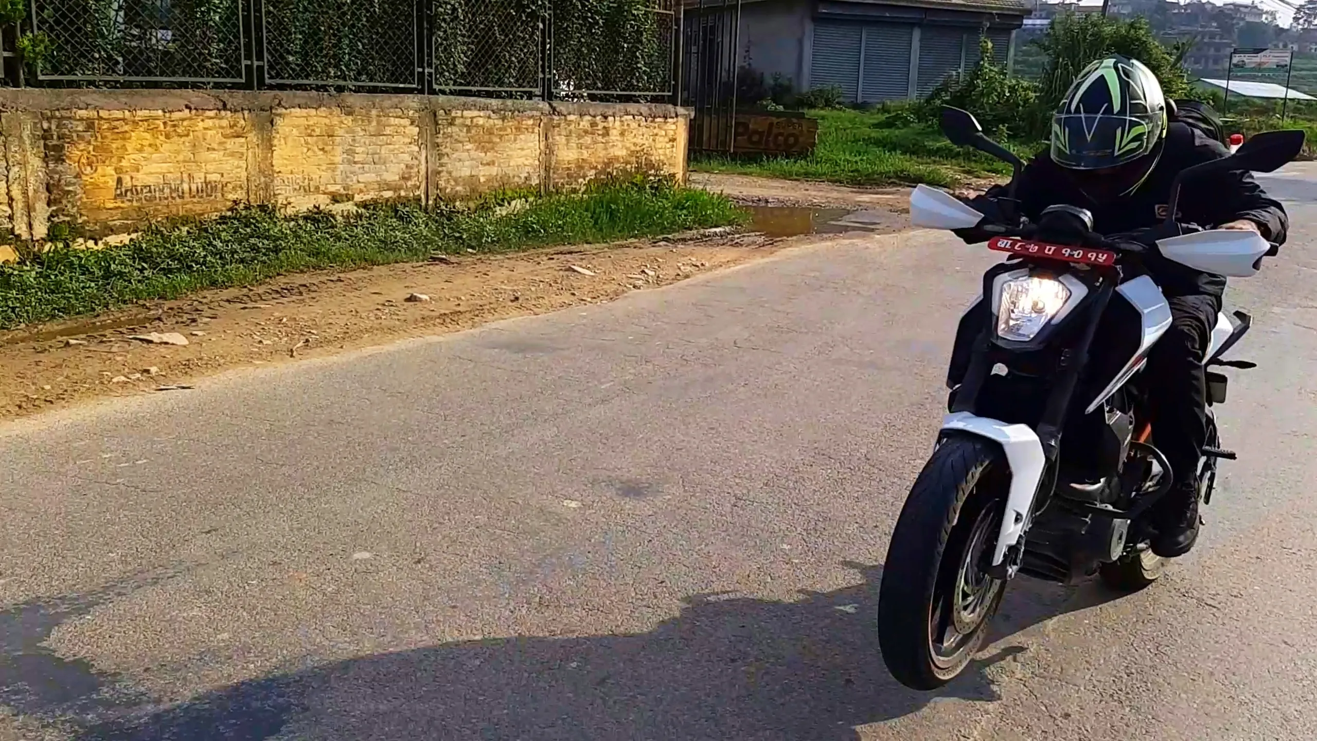 Action shot of a motorcyclist riding a white and black KTM Duke style naked sportbike with sharp angular headlight design along a quiet paved rural road in the Kathmandu Valley, Nepal. The rider wears a full-face helmet with green and white graphic design, black riding jacket, and black gloves, with a passenger seated behind also dressed in black riding gear. The bike has a red Nepali number plate visible on the front fender. Background shows a stone and brick boundary wall with green ivy growing on a metal fence, lush green grass, a small shop with shuttered doors, and distant hills with scattered houses under a hazy sky. This captures the thrill of motorbike touring through Nepal's countryside roads, Kathmandu to Bethanchowk bike ride, adventure motorcycling Nepal, KTM Duke touring Himalayas.