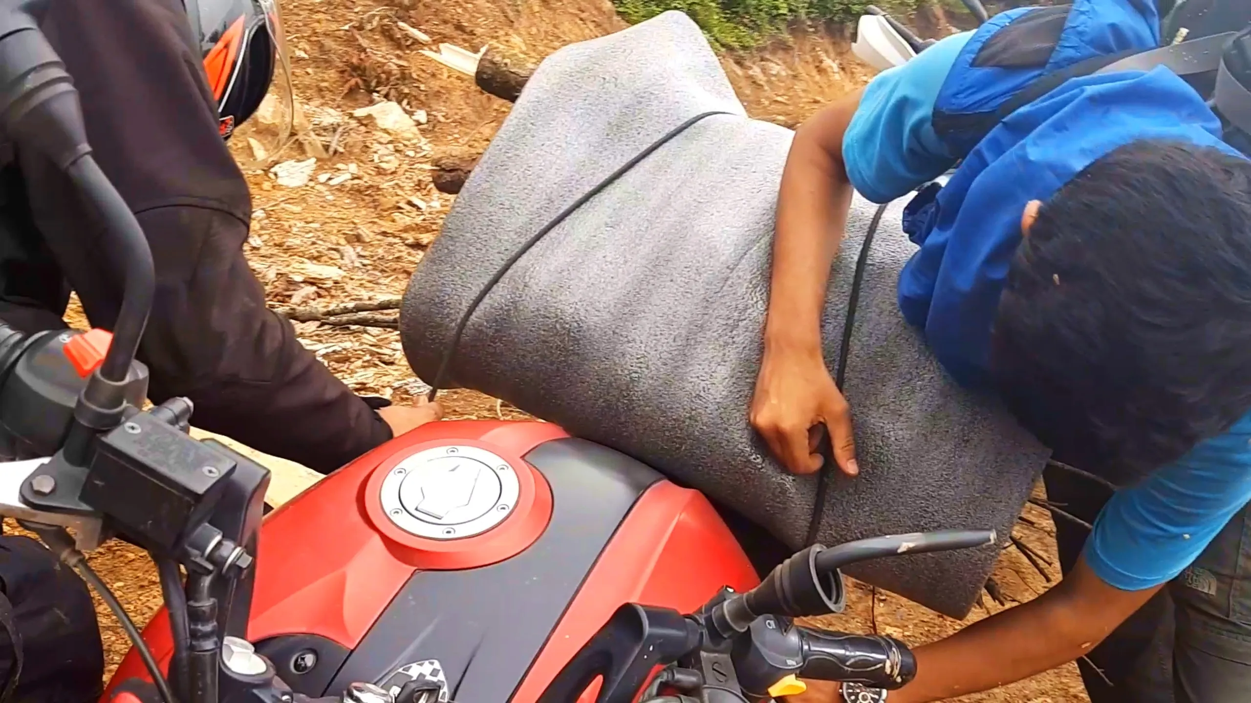 Close-up shot of two hands working together to pack a thick rolled grey sleeping mat or foam mattress onto the rear of a red and black motorbike, with a black bungee cord securing the bundle. One person wearing a black riding jacket and helmet stands on the left, while another in a sky-blue t-shirt and blue rain jacket reaches across from the right, both hands gripping the rolled mat to tighten it onto the bike's red fuel tank and seat. The dirt ground scattered with twigs and rocks is visible in the background near dense green forest. This shows packing camping gear onto motorbikes for Bethanchowk overnight trip, Nepal motorbike trekking preparation, Himalayan adventure gear setup, group camping logistics Kathmandu hills.