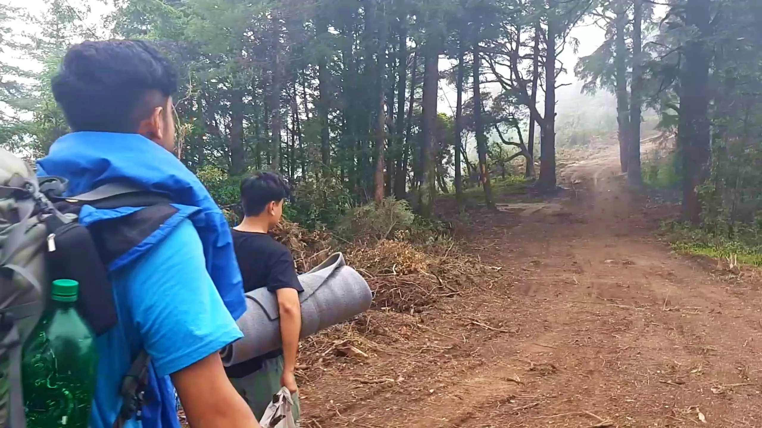 Photo of two young hikers walking away from the camera along a misty pine forest dirt trail strewn with dry pine needles and fallen branches. The hiker in front wears a bright blue rain jacket draped over his shoulders covering a backpack, with a green plastic water bottle visible at his side, while his friend ahead carries a long grey rolled sleeping mat across his back, wearing a black t-shirt and green cargo shorts. Tall pine and cedar trees with hazy fog create an atmospheric tunnel-like view of the trail leading toward a clearing in the distance. This captures the scenic forest trekking trail at Bethanchowk, Nepal pine forest hiking adventure, Himalayan misty trail trekking, backpacking camping gear Kathmandu hills.