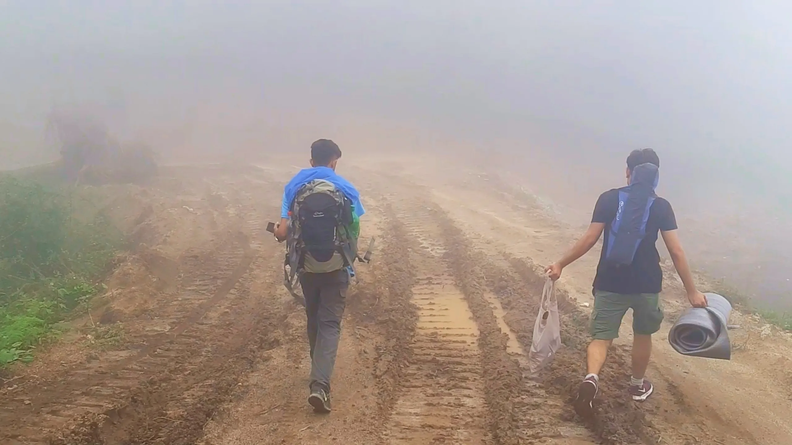 Photo taken from behind of two hikers walking along a muddy tire-rutted dirt road completely enveloped in thick white fog with zero visibility of surroundings. The hiker on the left wears a large green and black hiking backpack with a sky-blue rain jacket draped over the top, walking with hiking poles or a phone in hand. The hiker on the right carries a long grey rolled sleeping mat across his back and a white plastic bag in his hand, wearing a black t-shirt and green cargo shorts with red and white sneakers. Deep muddy tire tracks crisscross the orange-brown dirt road disappearing into the dense fog ahead. This captures the extreme foggy conditions on Bethanchowk muddy trekking trail, Nepal monsoon hiking adventure, Himalayan whiteout fog trekking, off-road camping trip Kathmandu hills.