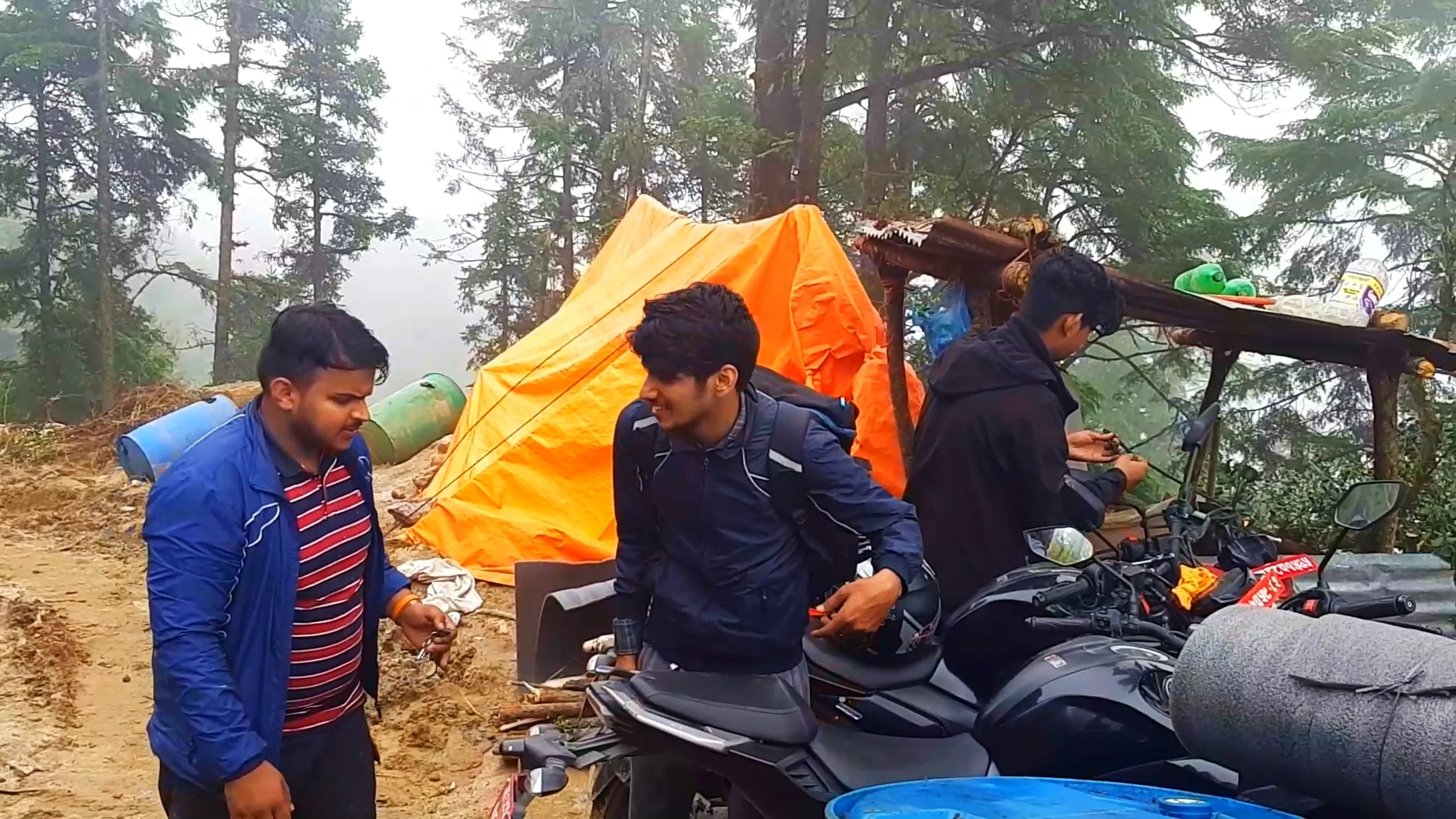 Group photo of three young men setting up camp near a bright orange triangular tent pitched among tall pine and cedar trees on a misty mountain campsite. The man in the foreground wears a blue jacket with red and navy striped shirt, holding a small object, smiling at the camera. Behind him, another man in a navy blue jacket with a grey backpack stands near several motorbikes including one with rolled sleeping mats strapped to the back, while a third man in a black jacket works on motorbike equipment. Blue plastic barrels, green water bottles, firewood logs, and a makeshift wooden shelf structure are visible around the campsite. Dense fog-covered pine forest surrounds the clearing. This shows the overnight camping setup at Bethanchowk highest point, Nepal jungle camping tent site, Himalayan motorbike trekking campsite, group adventure camping Kathmandu hills.