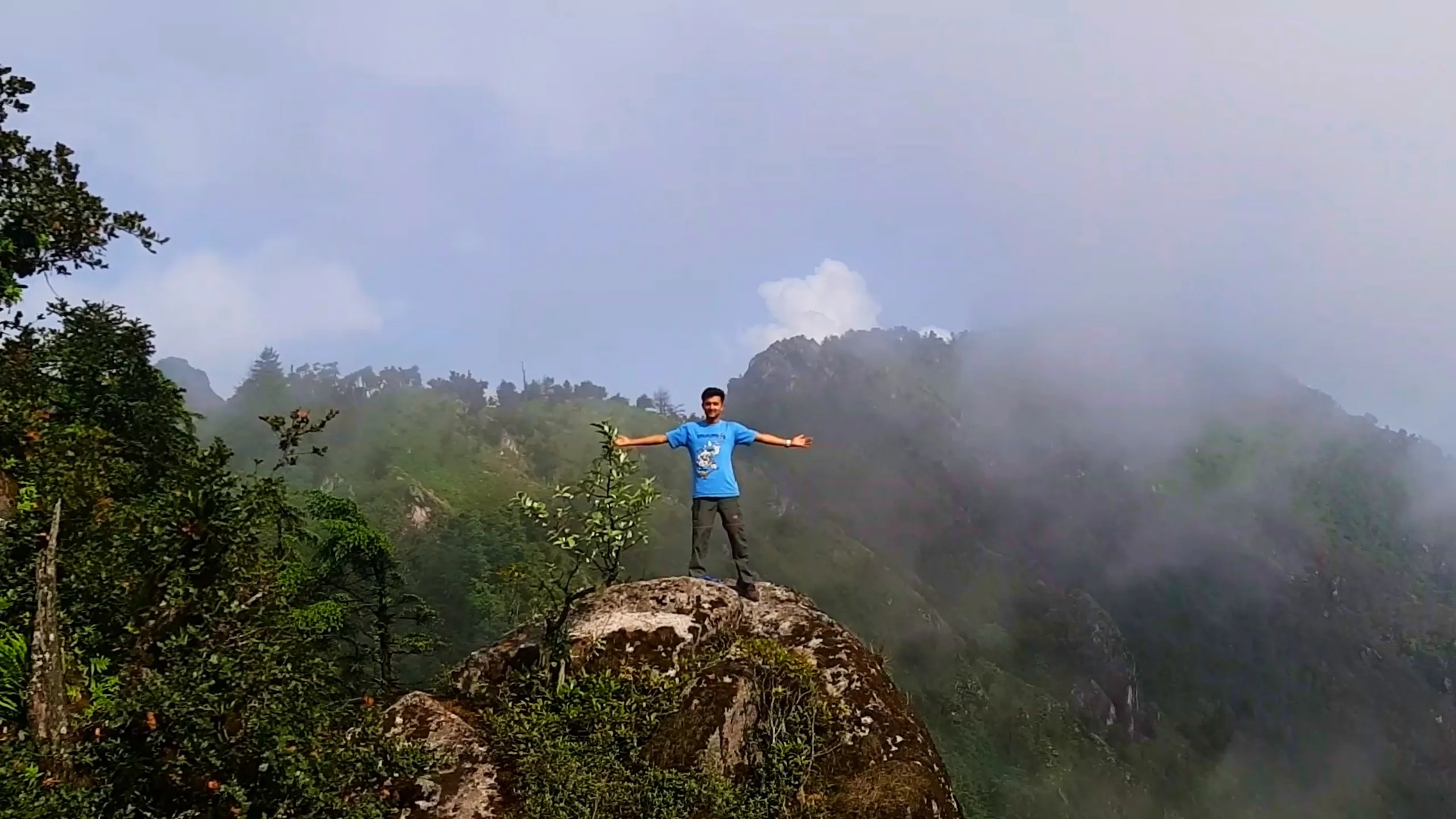 Triumphant photo of a young man wearing a sky-blue "Wish List Nepal" graphic t-shirt and grey cargo pants standing with arms spread wide on top of a large moss-covered boulder at a misty mountain viewpoint, smiling confidently at the camera. Behind him, layered green forested hills and rocky ridges fade into thick white clouds and fog covering the distant peaks. Small shrubs and trees frame the boulder on the left side. This captures the iconic summit celebration pose at Bethanchowk highest point, Nepal mountain peak achievement, Himalayan trekking summit photography, adventure travel milestone Kathmandu hills.