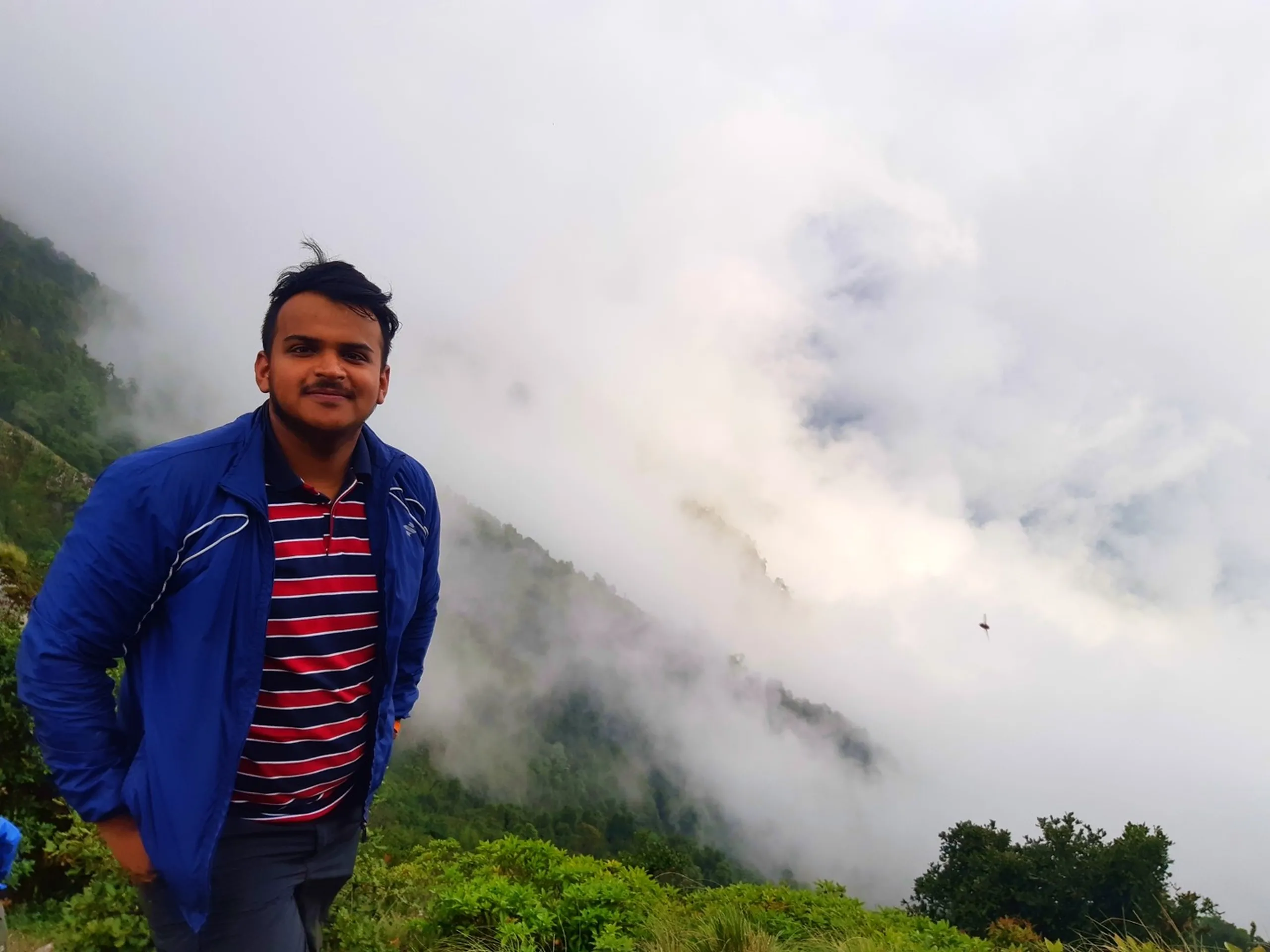 Portrait photo of a smiling young man with short dark hair wearing a blue zip-up jacket over a red and navy striped polo shirt, standing on a grassy hillside with one hand in his pocket. Behind him, a dramatic backdrop of thick white clouds and mist rolls over green forested mountain slopes, with a small bird visible flying through the misty sky. Lush green shrubs and grass surround him in the foreground. This captures a scenic portrait at Bethanchowk misty mountain viewpoint, Nepal Himalayan adventure travel photography, foggy mountain trekking portrait, group hiking trip memories Kathmandu hills.