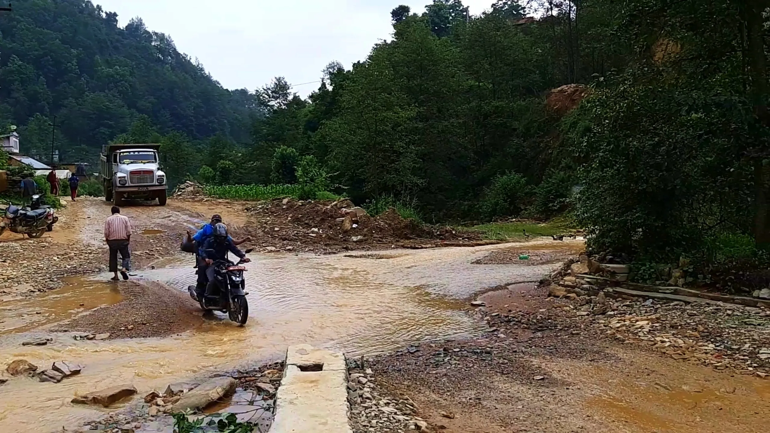 Documentary-style photo of a motorbike with two riders wearing helmets and a blue raincoat-wrapped backpack crossing a muddy flooded dirt road during monsoon season in rural Nepal, surrounded by dense green forested hills. A white dump truck is parked ahead on the muddy track near small village houses, while a pedestrian in a striped shirt and slippers walks alongside, and another parked motorbike rests near the roadside. Reddish-brown muddy water floods across the rocky unpaved road, with construction debris and stones scattered around, reflecting the challenging monsoon road conditions typical of off-road trekking routes to Bethanchowk Kathmandu, rural Nepal road trip, monsoon trekking Nepal, off-road motorbike adventure Himalayas.