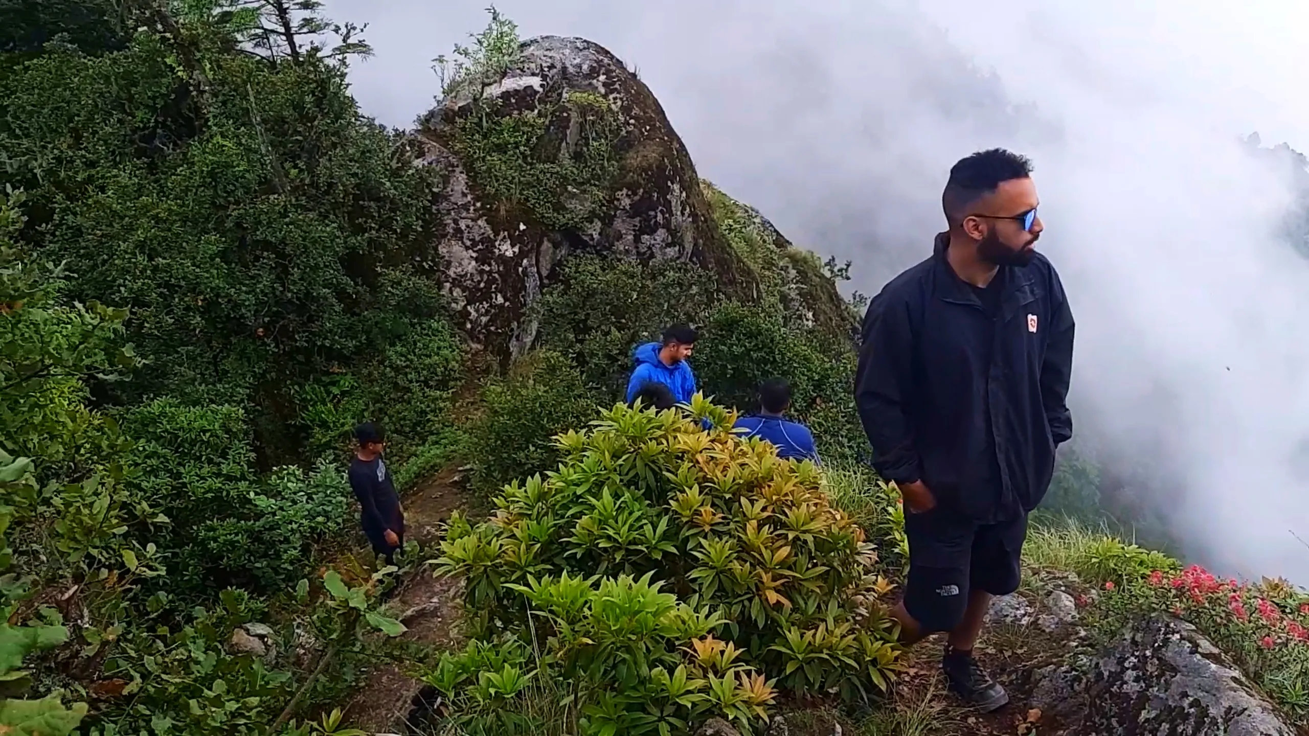 Group photo of young male hikers walking along a narrow ridge trail at Bethanchowk highest point, surrounded by dense green rhododendron shrubs and moss-covered boulders with blooming pink flowers on the right edge. In the foreground, a man wearing mirrored sunglasses, a dark navy windbreaker jacket with red logo patch, and black North Face shorts walks confidently with hands in pockets, looking off to the side. Behind him on the trail, other friends in blue jackets and dark t-shirts sit and stand among the bushes near a large lichen-covered rock formation. Thick white clouds and fog roll dramatically over the cliffside in the background. This captures the scenic ridge trail hiking experience at Bethanchowk summit, Nepal Himalayan trekking adventure, misty mountain ridge walk Kathmandu hills, group hiking trip rhododendron forest.