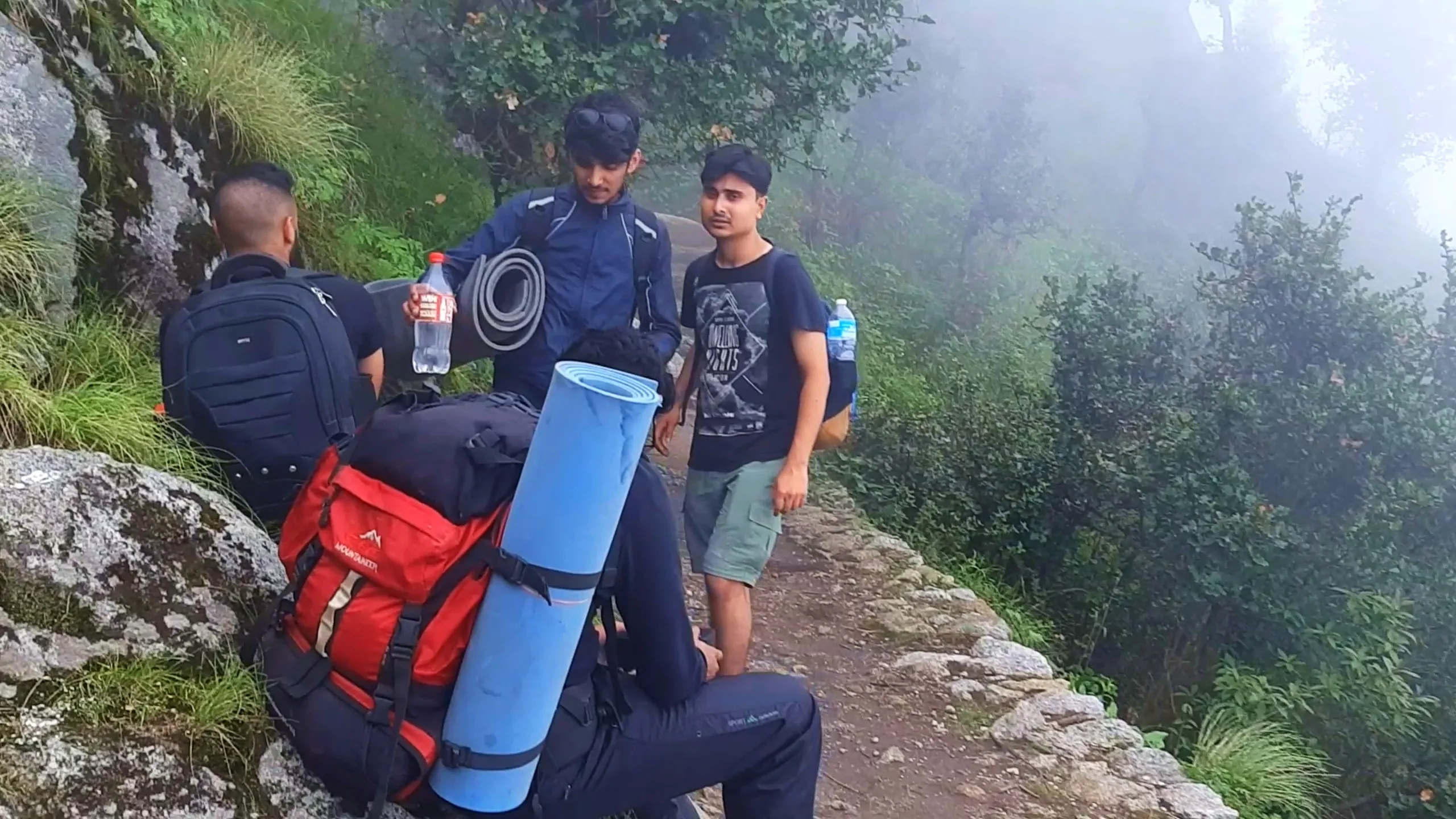 Group photo of four young male hikers resting on a narrow ridge trail at Bethanchowk, surrounded by misty green rhododendron bushes and large grey boulders. Two friends sit on large camping backpacks, one bright red "Mountainer" branded backpack with a blue rolled sleeping mat strapped on top, and another black backpack, while a man in a navy blue jacket with sunglasses on his head holds a rolled grey sleeping mat and a water bottle with red label. Another friend in a black "Amazing Nepal" graphic t-shirt and green cargo shorts stands nearby holding a blue water bottle. Thick white fog blankets the forested valley below. This captures a rest stop with camping gear during Bethanchowk trekking trip, Nepal hiking backpack essentials, Himalayan misty mountain trail break, group adventure camping gear Kathmandu hills.