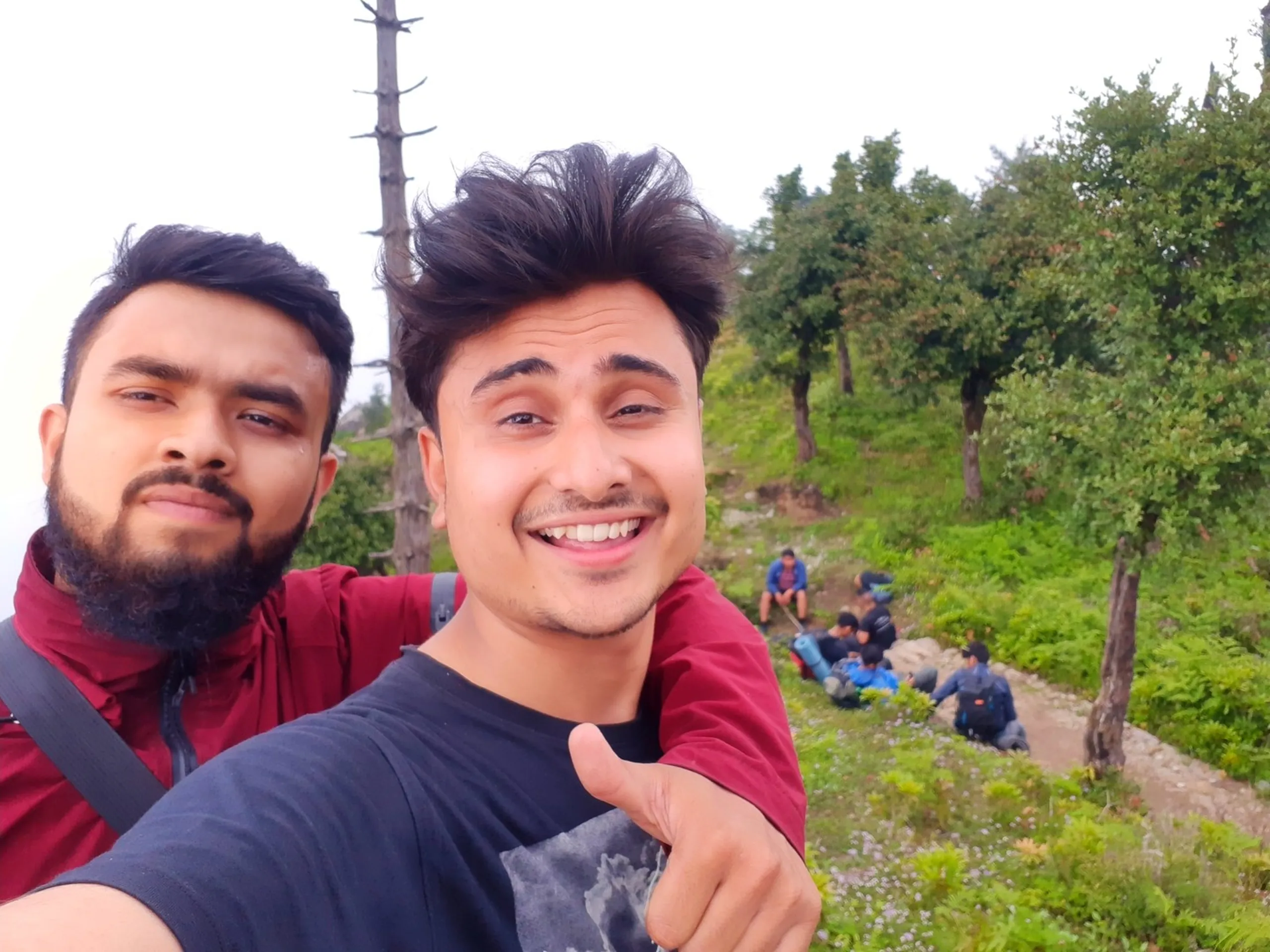 Cheerful close-up selfie of two young men at a misty hilltop viewpoint in Bethanchowk, Nepal. The man in front has voluminous styled dark hair, wearing a navy blue t-shirt with a faded graphic design, giving a thumbs up gesture with a big smile. Behind him, a bearded friend wearing a maroon red rain jacket and a backpack with a grey strap looks at the camera with a neutral expression. In the background, blurred figures of other hikers sit and relax on a grassy hillside path lined with green shrubs and trees under an overcast white sky. This captures a joyful summit selfie moment at Bethanchowk highest point, Nepal Himalayan trekking friends, mountain peak adventure photography, group hiking trip memories Kathmandu hills.