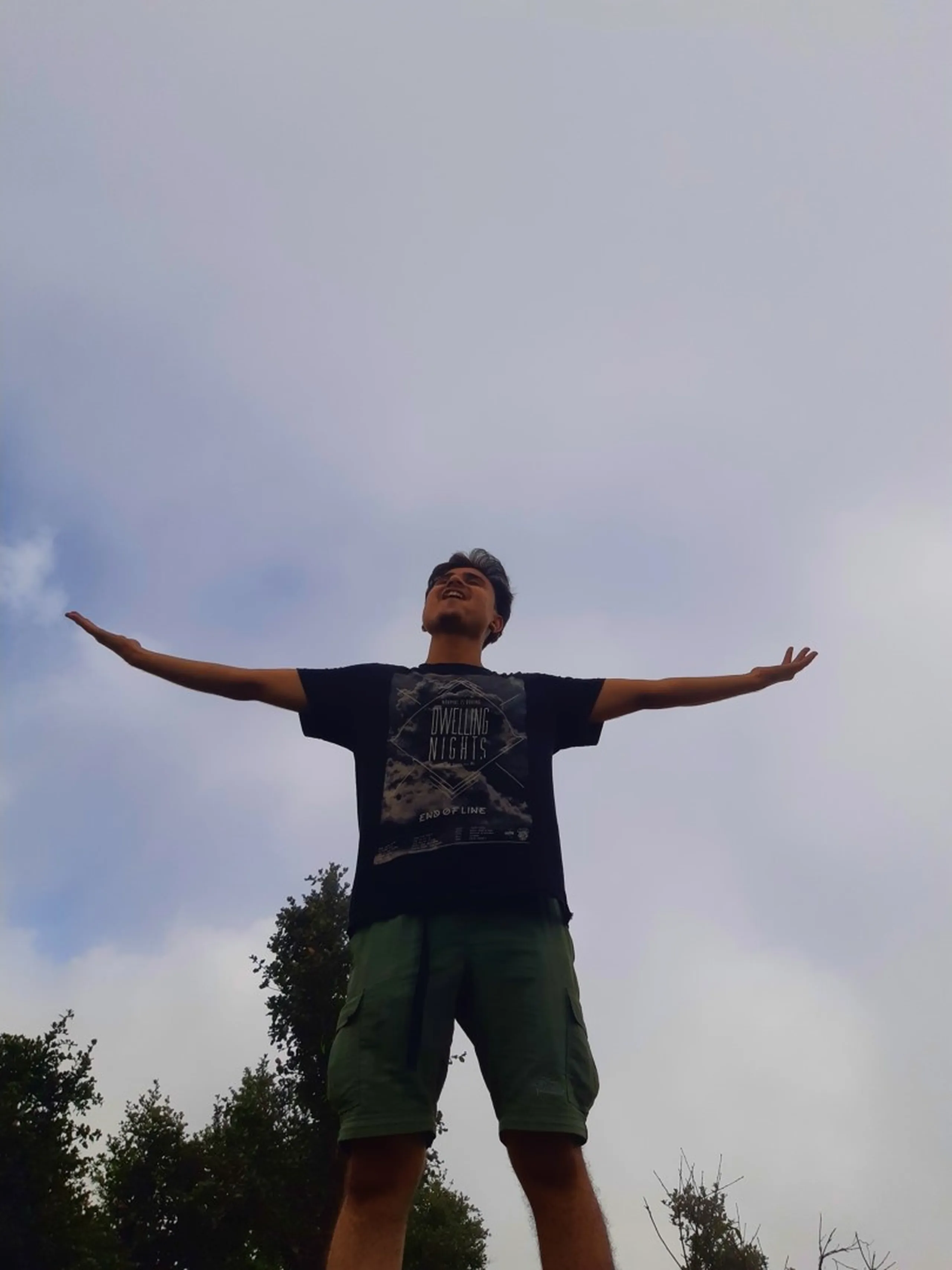 Triumphant low-angle photo of a young man standing with arms spread wide against an overcast cloudy sky, wearing a black "Dwelling Nights End of Line" graphic t-shirt and olive green cargo shorts, looking upward with a joyful expression. Green tree branches are visible at the bottom edges of the frame against the bright grey-white sky. This captures a free-spirited celebration pose at Bethanchowk mountain summit, Nepal adventure travel photography, Himalayan trekking achievement moment, scenic hilltop pose Kathmandu hills.