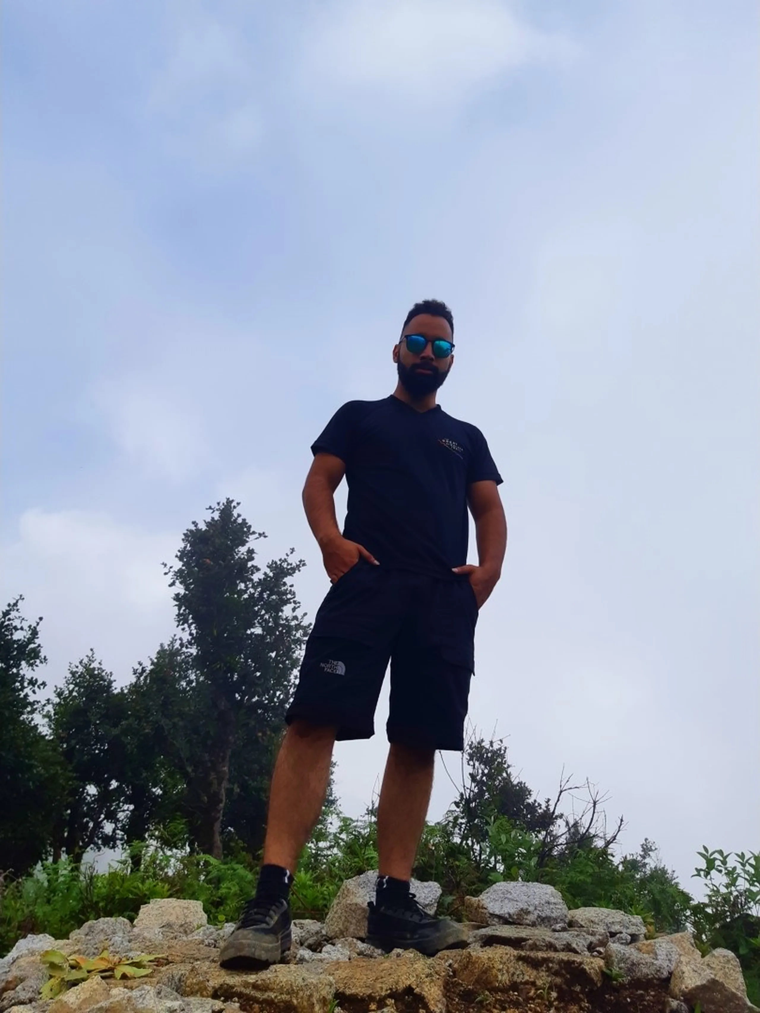 Confident low-angle portrait of a bearded young man standing on a pile of grey rocks at a misty hilltop, wearing a black "The North Face" branded t-shirt and matching black cargo shorts with mirrored sunglasses, hands tucked into his pockets, looking directly at the camera with a serious expression. Green trees and shrubs are visible behind him against an overcast white and grey cloudy sky. This captures a stylish summit portrait at Bethanchowk highest point, Nepal Himalayan trekking fashion, mountain peak photography, adventure travel portrait Kathmandu hills.
