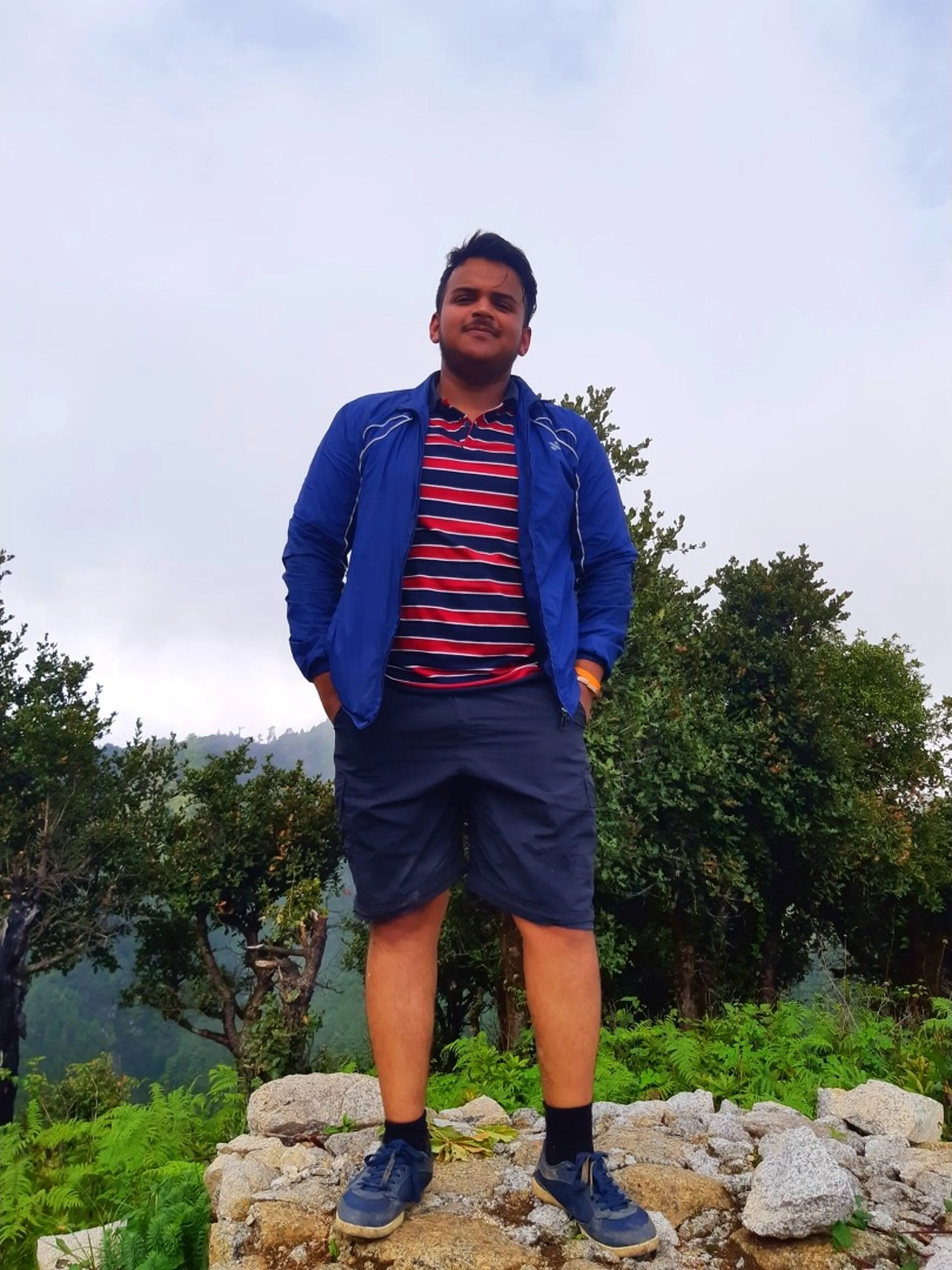 Full-body portrait of a young man standing confidently on a pile of grey rocks at a misty hilltop viewpoint in Bethanchowk, wearing a bright blue zip-up jacket over a red, navy, and white striped polo shirt with navy cargo shorts, blue sneakers, and an orange wristband, hands in pockets looking at the camera with a slight smile. Lush green ferns and trees frame him against an overcast white sky background. This captures a casual summit portrait during Bethanchowk trekking trip, Nepal Himalayan adventure travel fashion, mountain hiking photography, scenic hilltop pose Kathmandu hills.