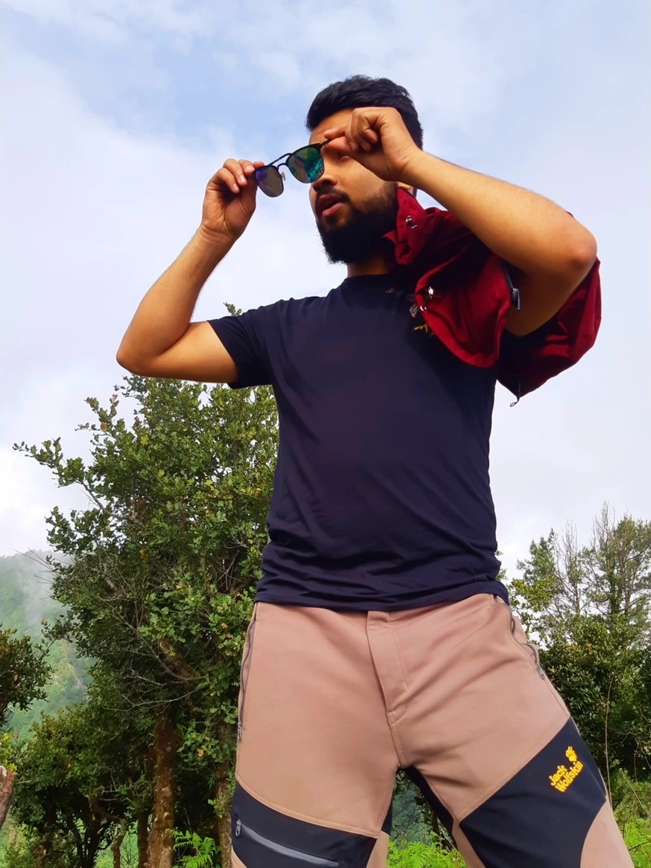 Dynamic low-angle action shot of a young bearded man adjusting his mirrored sunglasses with both hands, wearing a dark navy athletic t-shirt and tan-and-black "Jack Wolfskin" branded cargo pants, with a maroon red jacket draped over one shoulder. Green trees and misty hills are visible in the background against a bright overcast sky. This captures a stylish candid moment during Bethanchowk hiking trip, Nepal adventure travel fashion photography, Himalayan trekking gear showcase, outdoor lifestyle Kathmandu hills.
