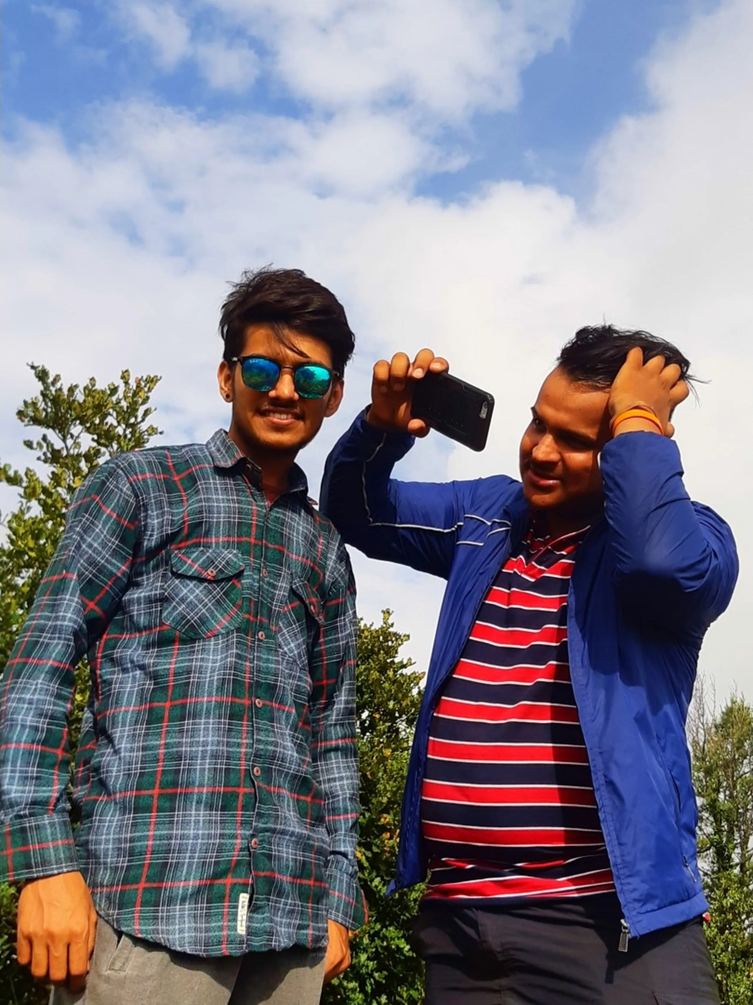 Candid photo of two young men taking a selfie together at a misty hilltop viewpoint in Bethanchowk. The man on the left wears a dark green and red checkered flannel shirt with mirrored sunglasses, smiling at the camera. Beside him, another man wearing a bright blue zip-up jacket over a red and navy striped polo shirt holds up a smartphone to take the photo while running his other hand through his hair, smiling. Green trees and bright white clouds fill the background. This captures a fun selfie moment during Bethanchowk trekking trip, Nepal hiking friends candid photography, Himalayan adventure travel memories, group selfie mountain viewpoint Kathmandu hills.