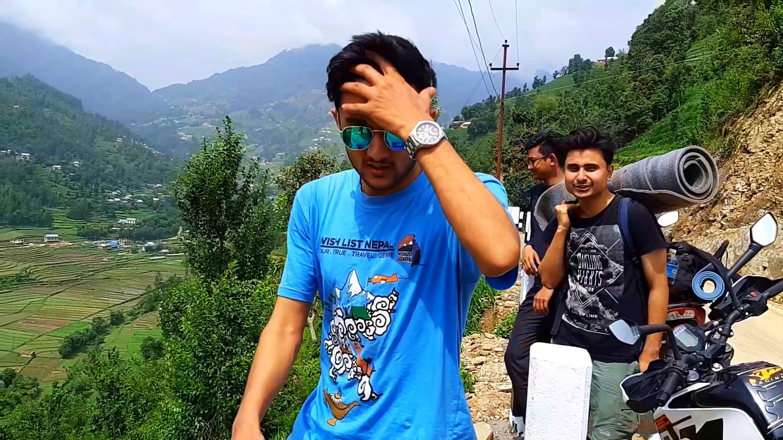 Candid travel photo of a young man in a sky-blue "Wish List Nepal" graphic t-shirt featuring a Himalayan mountain illustration with clouds, parachute, and lamp design, wearing mirrored aviator sunglasses and a silver wristwatch, brushing his hair back with his hand while standing on a hillside trekking trail. Behind him, terraced green rice paddy fields cascade down a misty valley with scattered village houses, and two other male hikers wearing backpacks (one carrying a rolled-up blue sleeping mat) walk along a paved mountain road lined with white concrete road markers, with a parked motorbike visible at the edge. Low clouds and fog roll over distant forested hills. This scene captures scenic Himalayan terrace farming landscape, Bethanchowk hiking trail views, Nepal countryside trekking photography, Wish List Nepal travel group adventure.