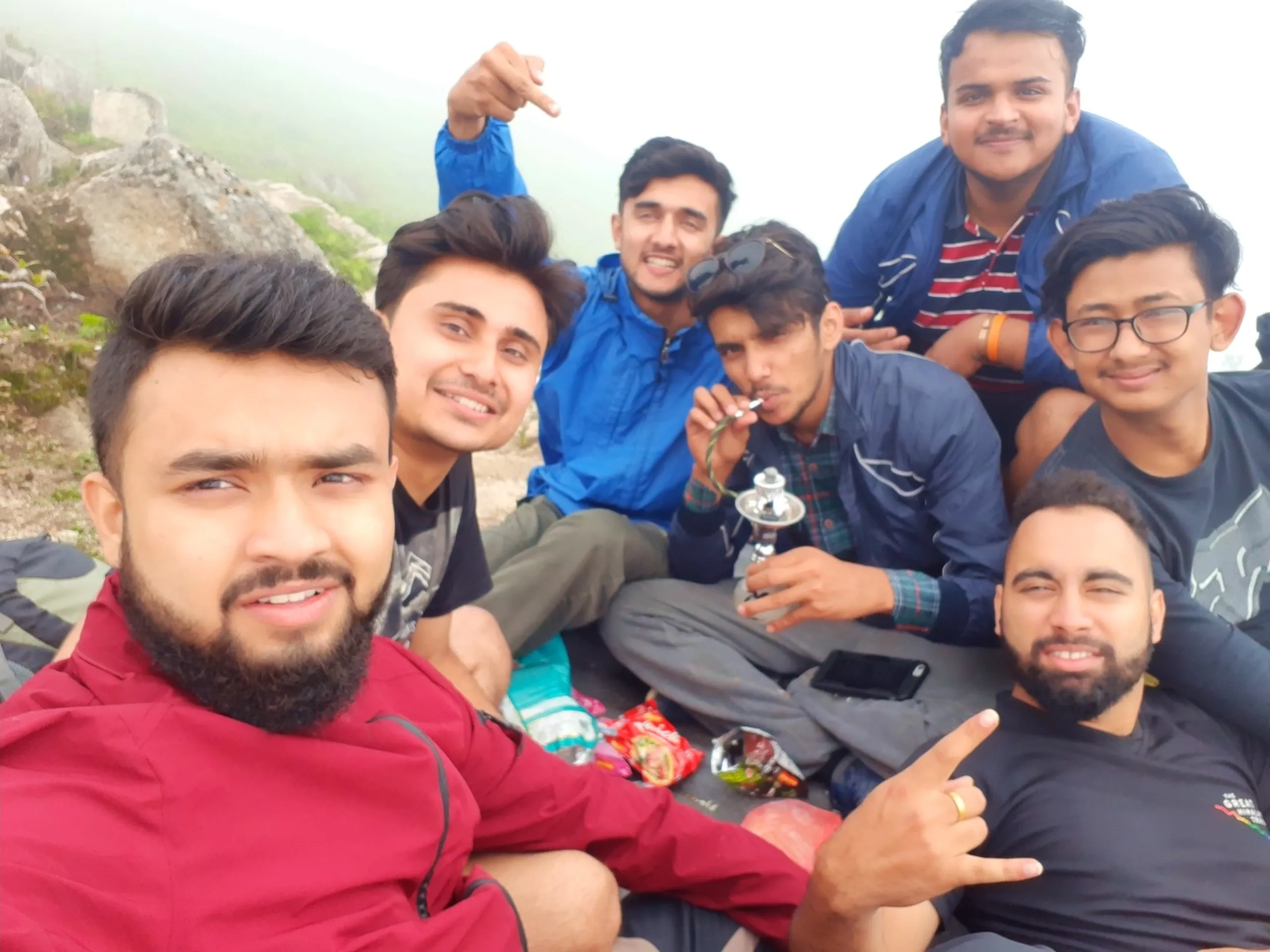 Joyful group selfie photo of seven young male friends sitting closely together at the foggy summit of Bethanchowk, Nepal, all smiling and posing for the camera. Various friends wear maroon red jackets, blue rain jackets, navy striped shirts, black t-shirts, and glasses, with one man in the center holding a small hookah or shisha pipe device to his mouth. Scattered snack wrappers, a water bottle, and a smartphone lie on the rocky ground between them. Thick white fog completely obscures any mountain view in the background, with grey rocks visible on the left edge. This captures the celebratory group summit moment at Bethanchowk highest point, Nepal trekking friends reunion, Himalayan group adventure photography, mountain peak celebration Kathmandu hills.