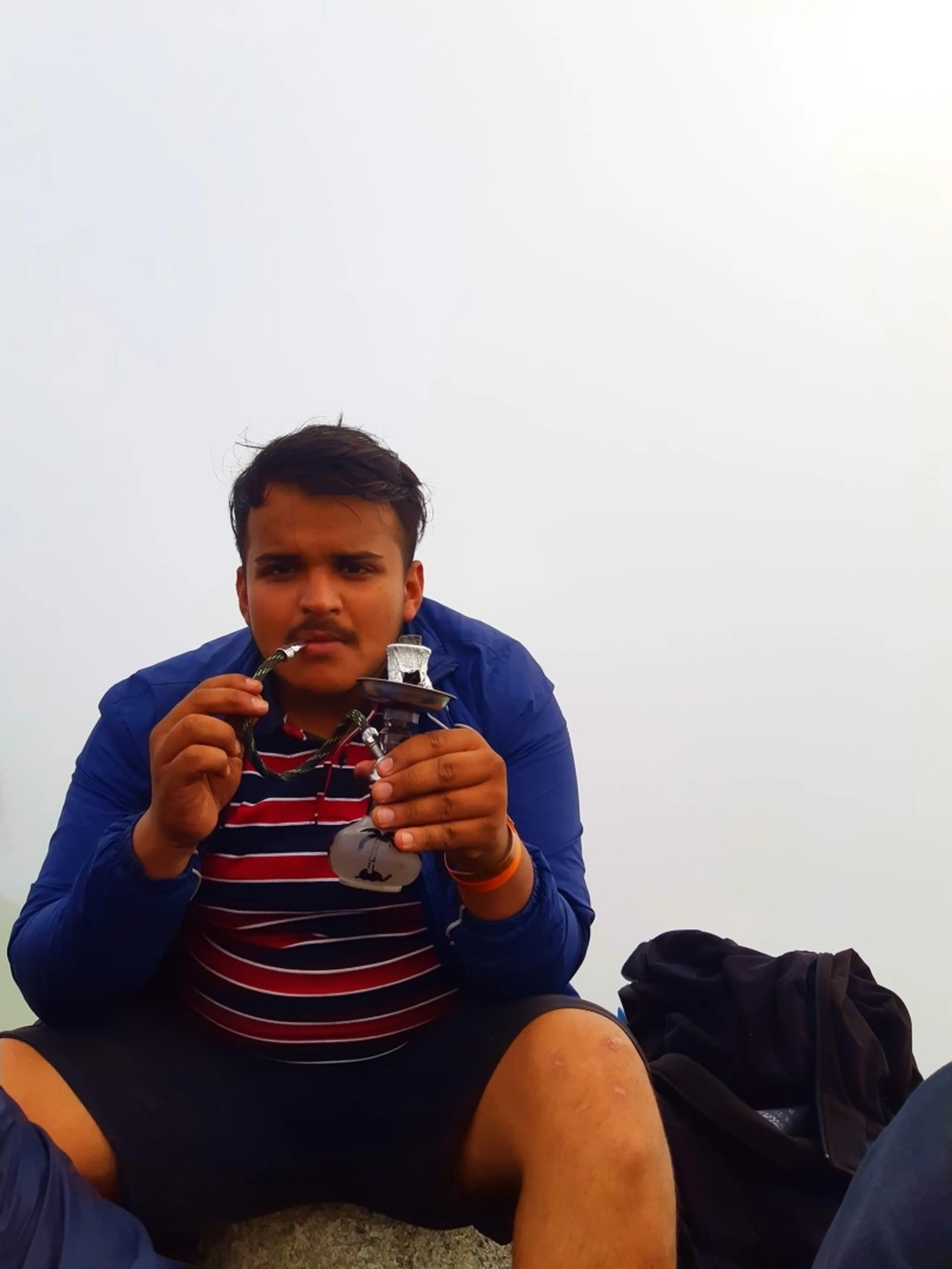Close-up portrait of a young man sitting cross-legged wearing a blue jacket over a red, navy, and white striped polo shirt with black shorts, holding a small portable hookah or shisha pipe device with a green hose to his mouth, wearing an orange wristband. A black jacket is bundled beside him on the rocky ground. Completely white foggy sky fills the entire background with no visible landscape. This captures a relaxed moment during Bethanchowk summit celebration, Nepal trekking group leisure activity, Himalayan misty mountain hangout, adventure trip relaxation Kathmandu hills.
