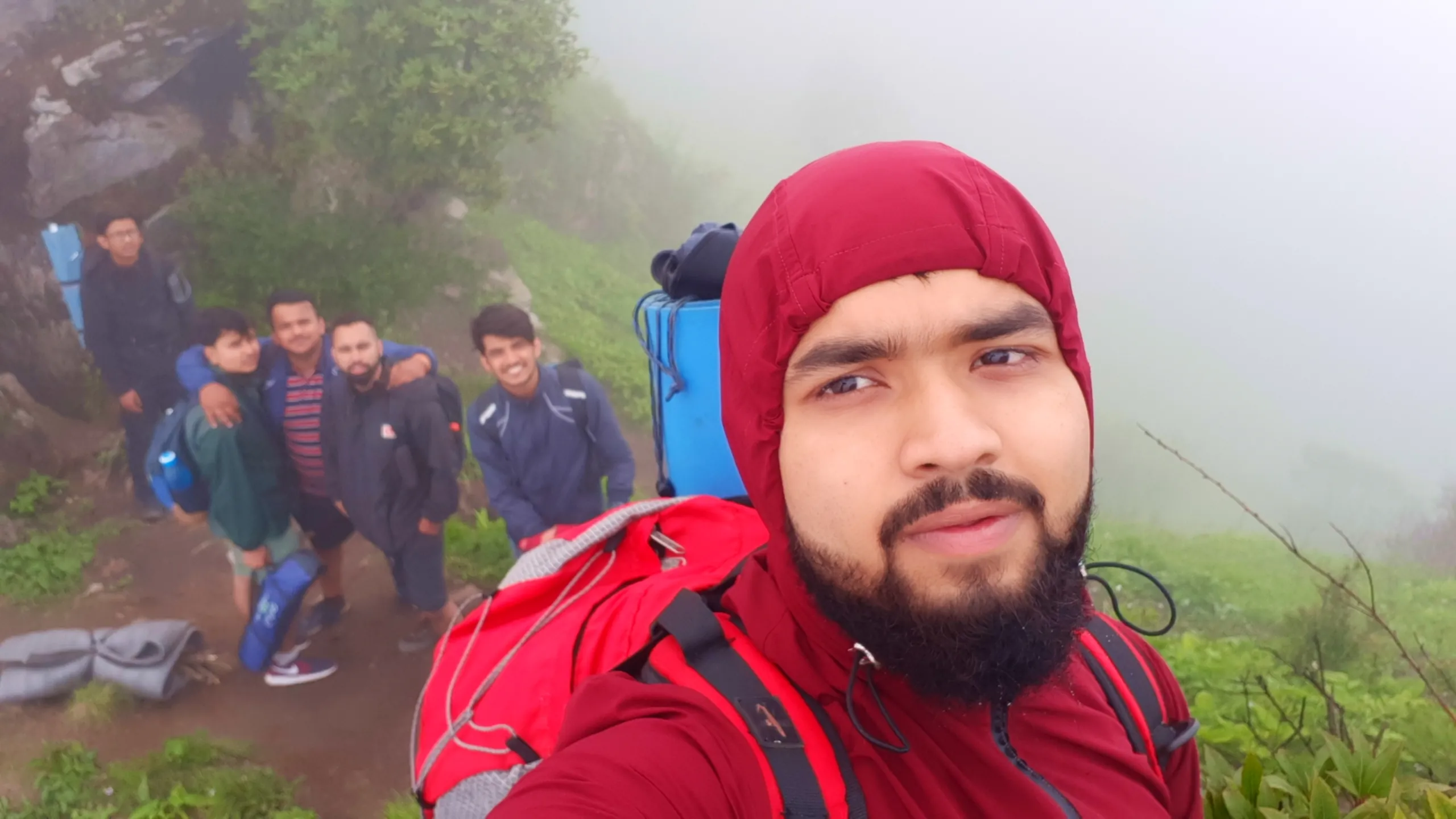 Close-up selfie of a bearded young man wearing a maroon red hooded rain jacket with the hood up, carrying a red "Mountainer" branded backpack with a blue rolled sleeping mat strapped on top, looking at the camera with a neutral expression. In the background, a group of blurred friends wearing blue and navy jackets with backpacks and rolled sleeping mats pose together on a misty grassy ridge trail near large grey boulders, with thick white fog covering the landscape. This captures a hooded selfie moment with the group during Bethanchowk trekking trip, Nepal Himalayan misty mountain hiking, adventure travel group photography, foggy ridge trail Kathmandu hills.